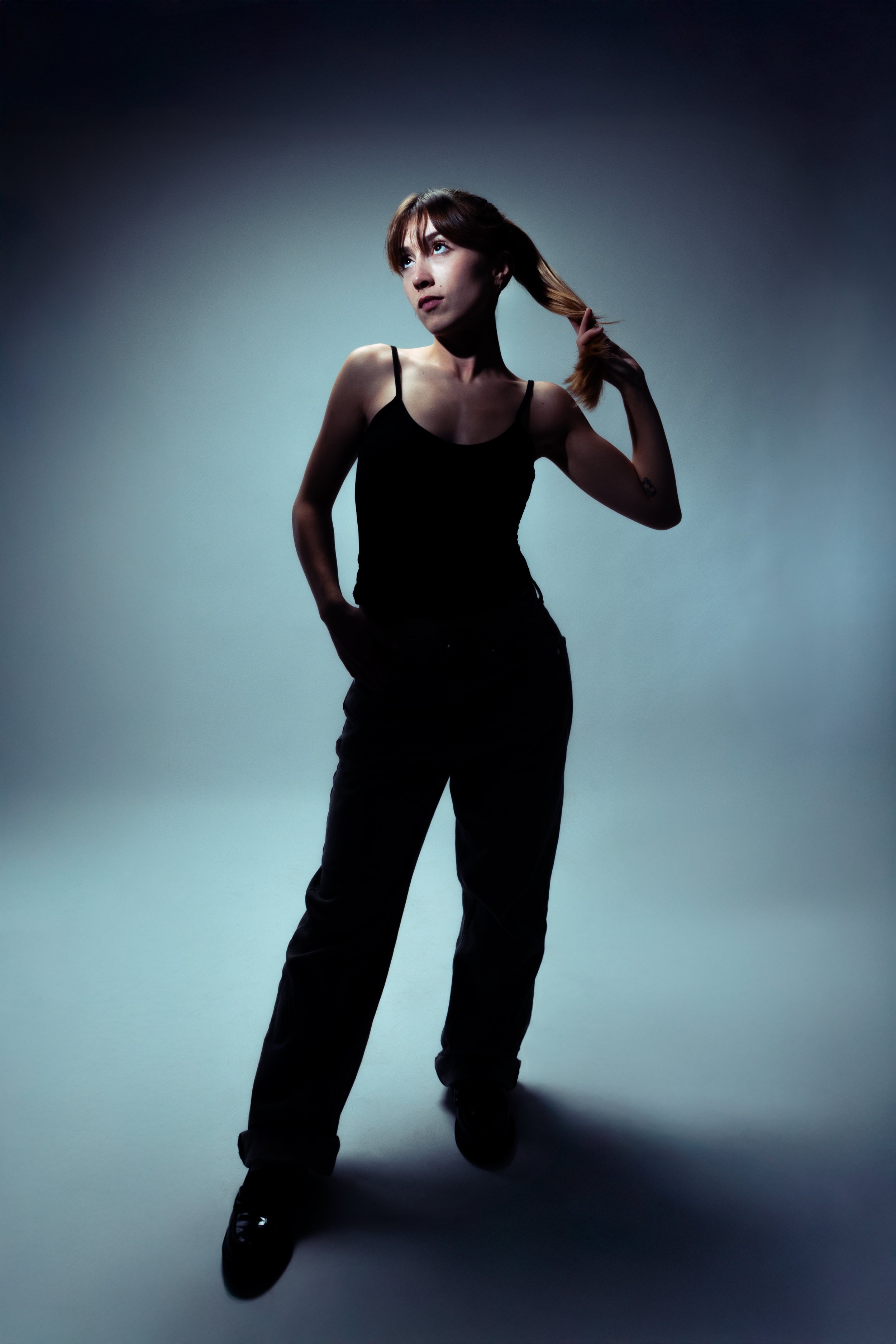 Une jeune femme debout, portant un débardeur noir et un pantalon noir, se coiffant avec ses cheveux longs et bruns, dans un environnement sombre avec un dégradé de lumière