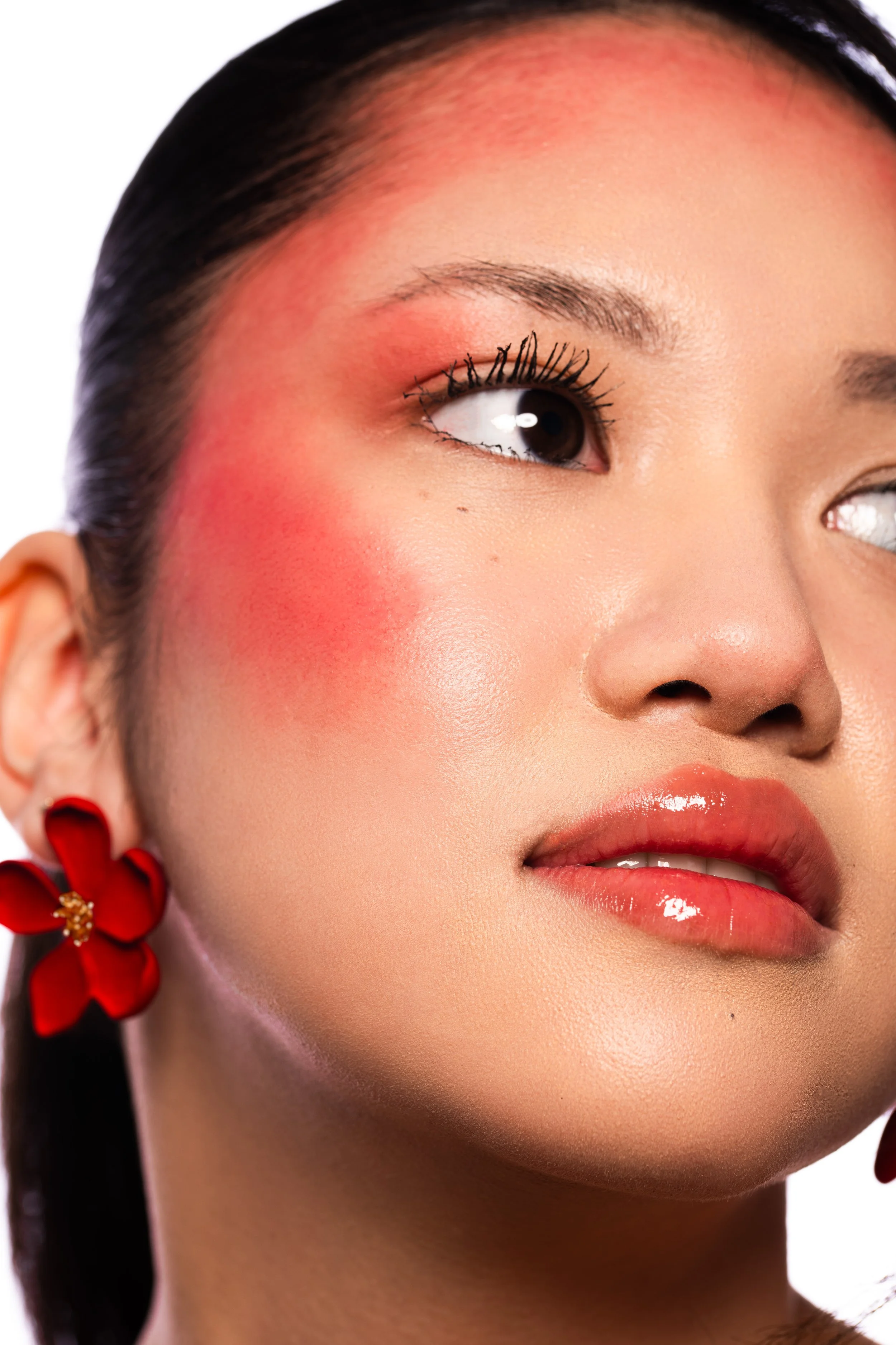 Close-up d'un visage de femme avec maquillage rose sur les joues, lèvres roses brillantes, oeil maquillé avec mascara, boucle d'oreille rouge en forme de fleur.