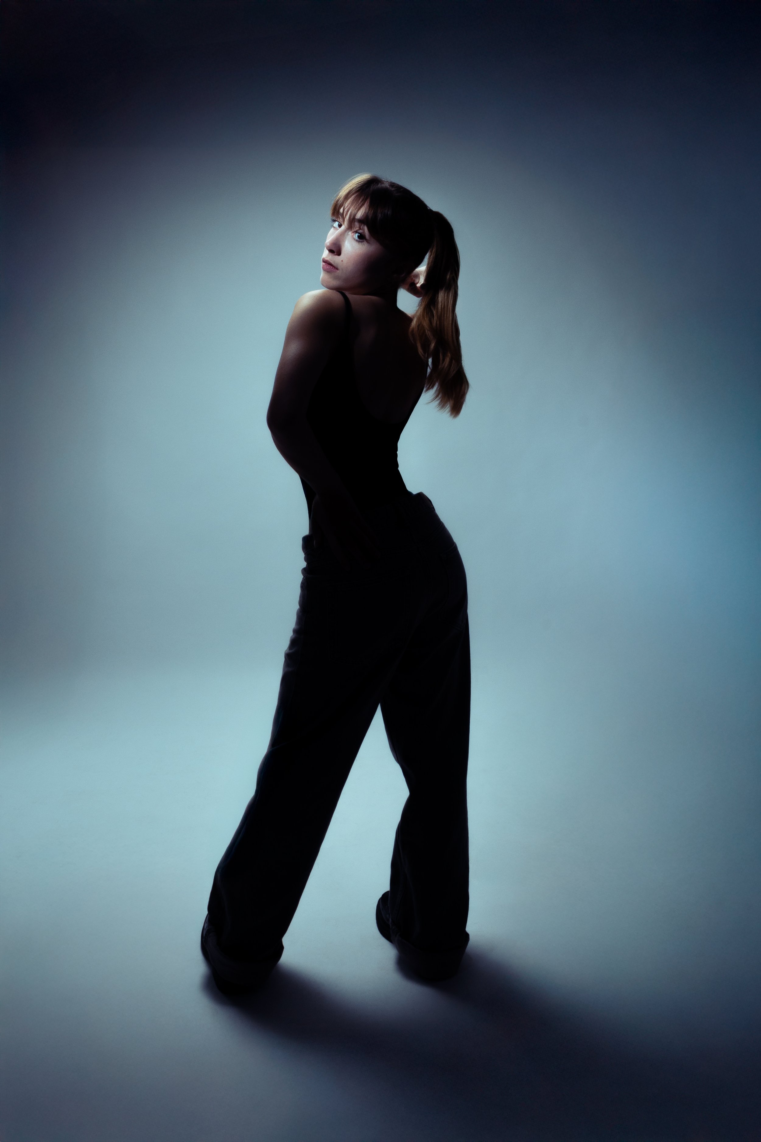 Une femme posant dans une lumière tamisée, portant une robe noire et des pantalons amples, vue de dos avec la tête tournée vers la caméra, fond neutre et ombragé.