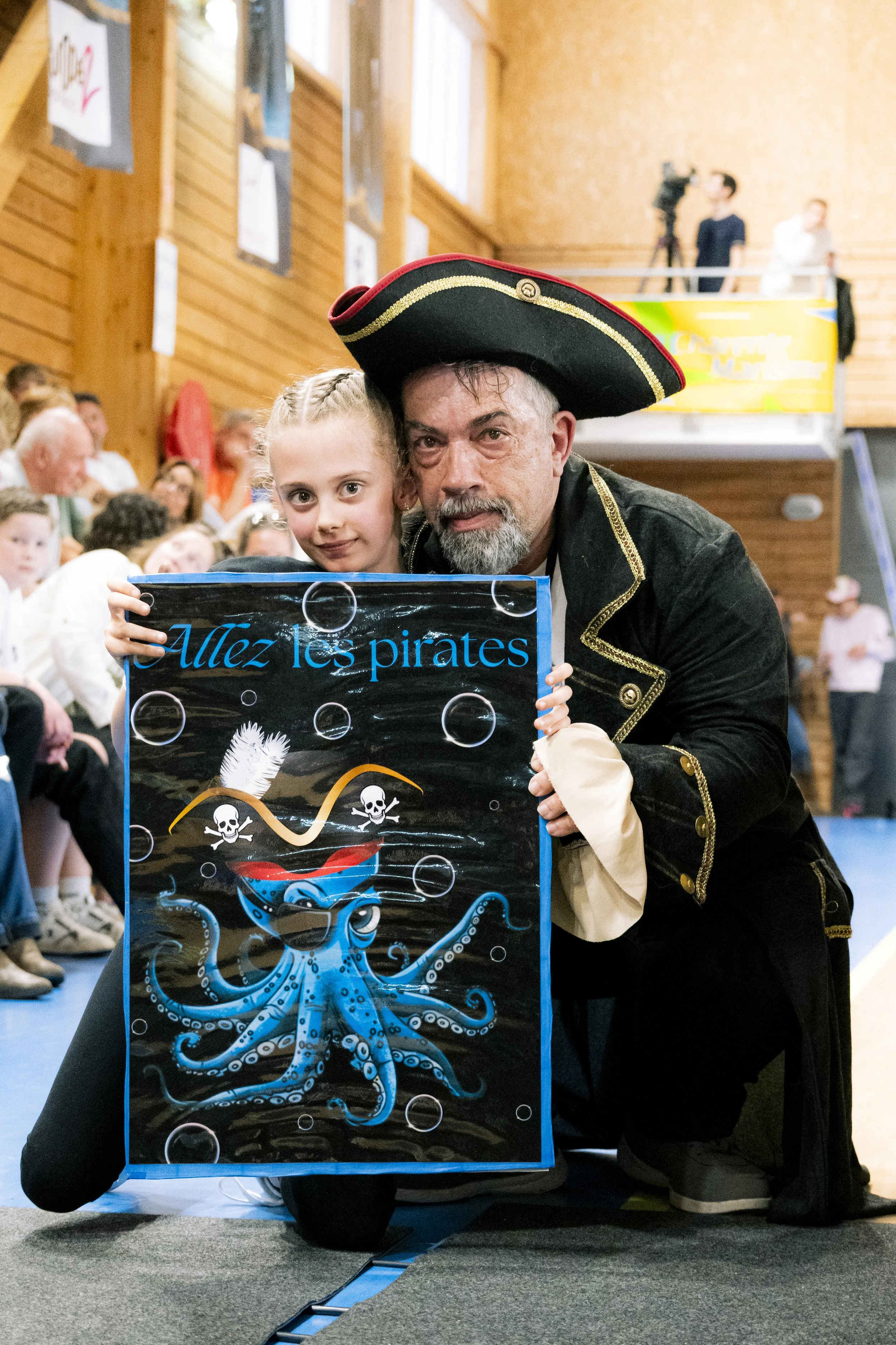Un homme et une jeune fille déguisés en pirates posent ensemble lors d'un événement, tenant une affiche avec un dessin d'un poulpe souriant portant un chapeau pirate, en arrière-plan un groupe de personnes assises dans une salle en bois.
