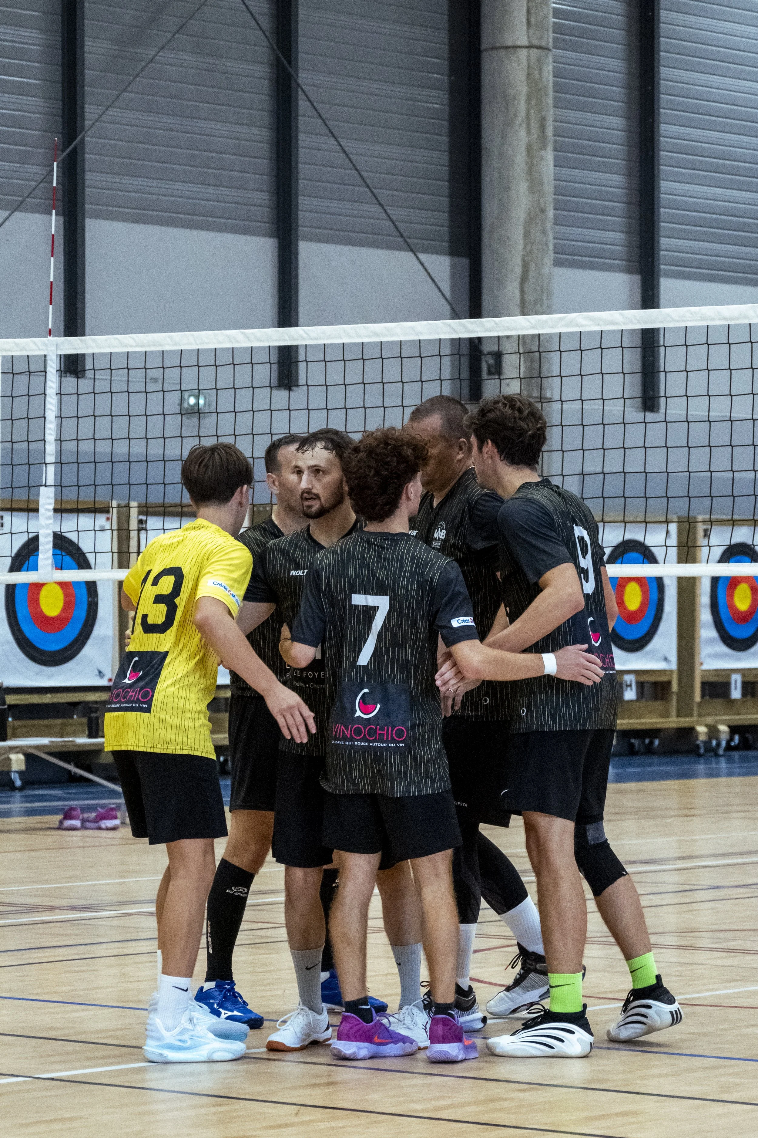 Un groupe de joueurs de volleyball discute sur un terrain intérieur, certains portant des maillots noirs et un en jaune, tous portant des chaussures de sport
