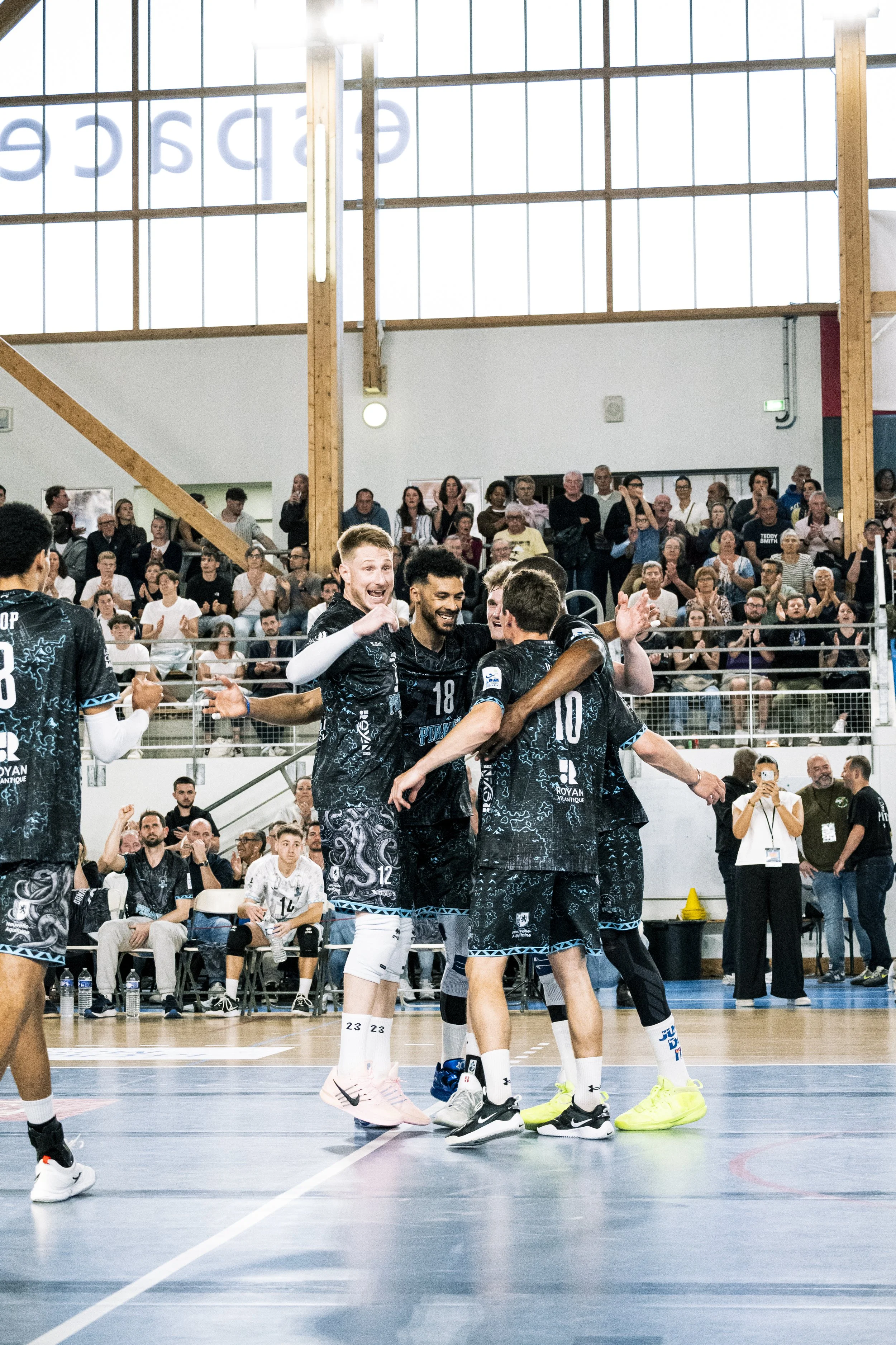 Des joueurs de volleyball en uniforme noir joyeux qui célèbrent un point sur le terrain intérieur avec un public enthousiaste en arrière-plan.