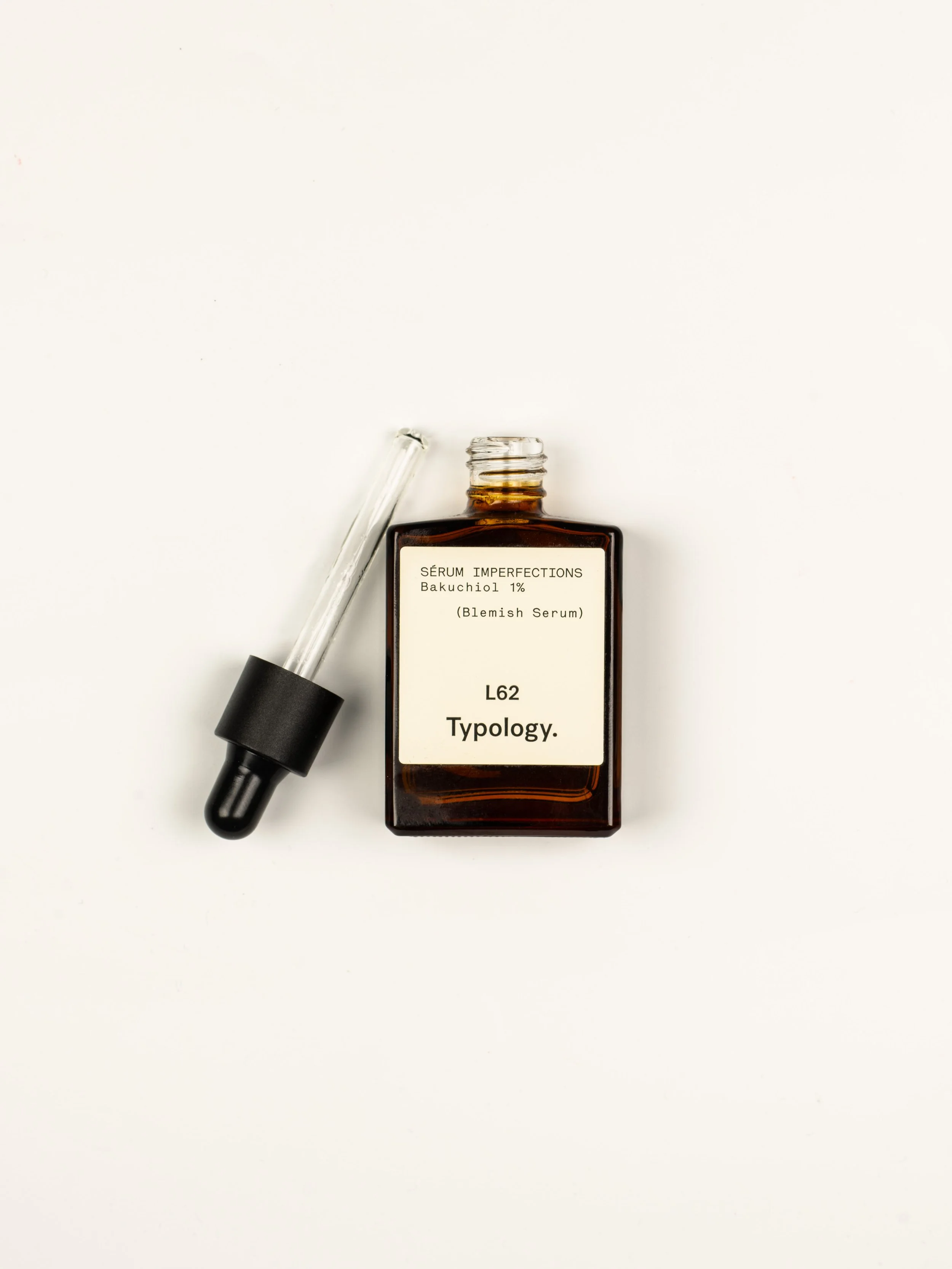 Un flacon brun portant une étiquette avec le texte 'SÉRUM IMPERFECTIONS Bakuchiol 1% (Blemish Serum) L62 Typology' et un pipette en verre à côté.