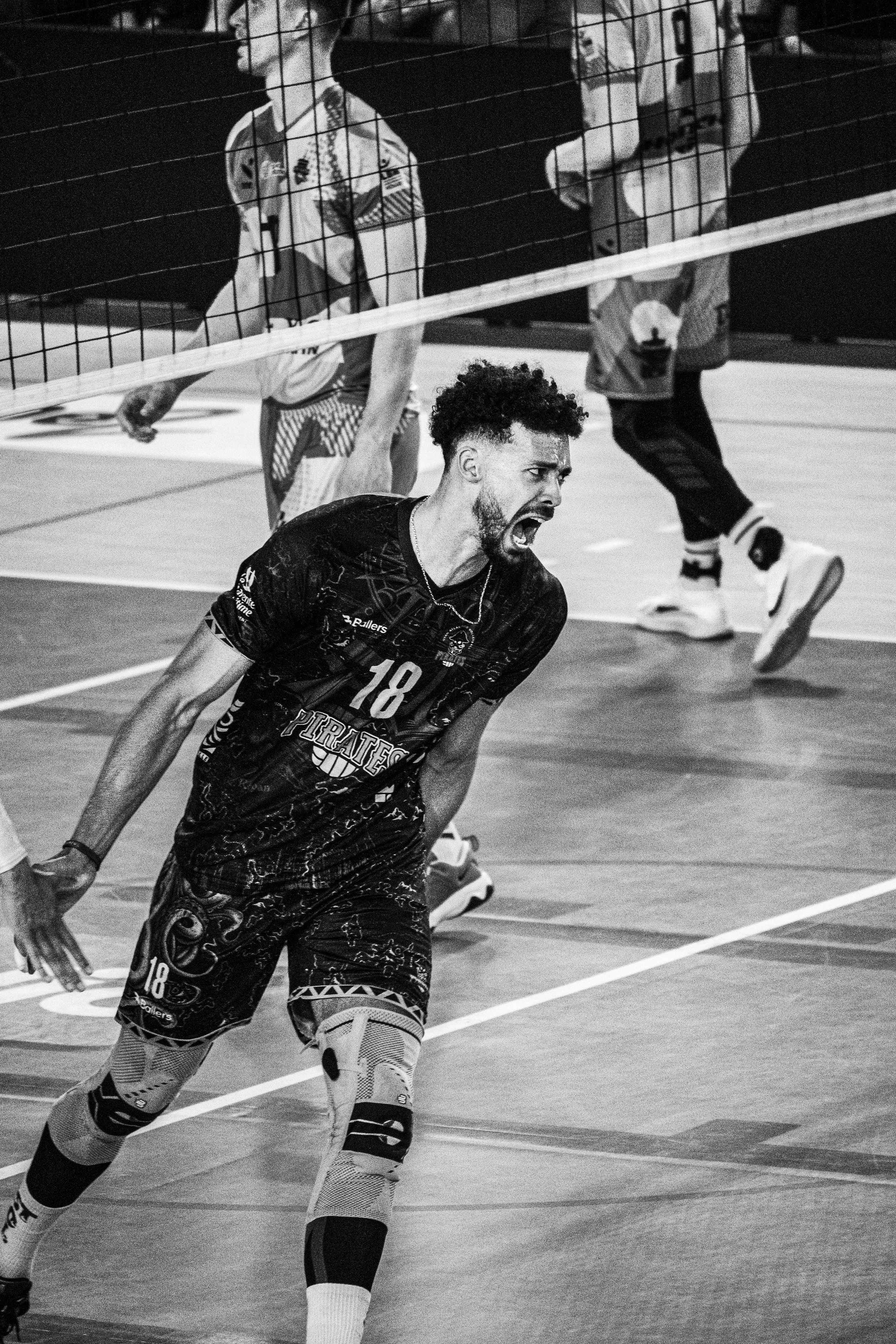 Un joueur de volley-ball en action lors d'un match, portant un maillot avec le numéro 18, sur un terrain intérieur avec des partenaires en arrière-plan.