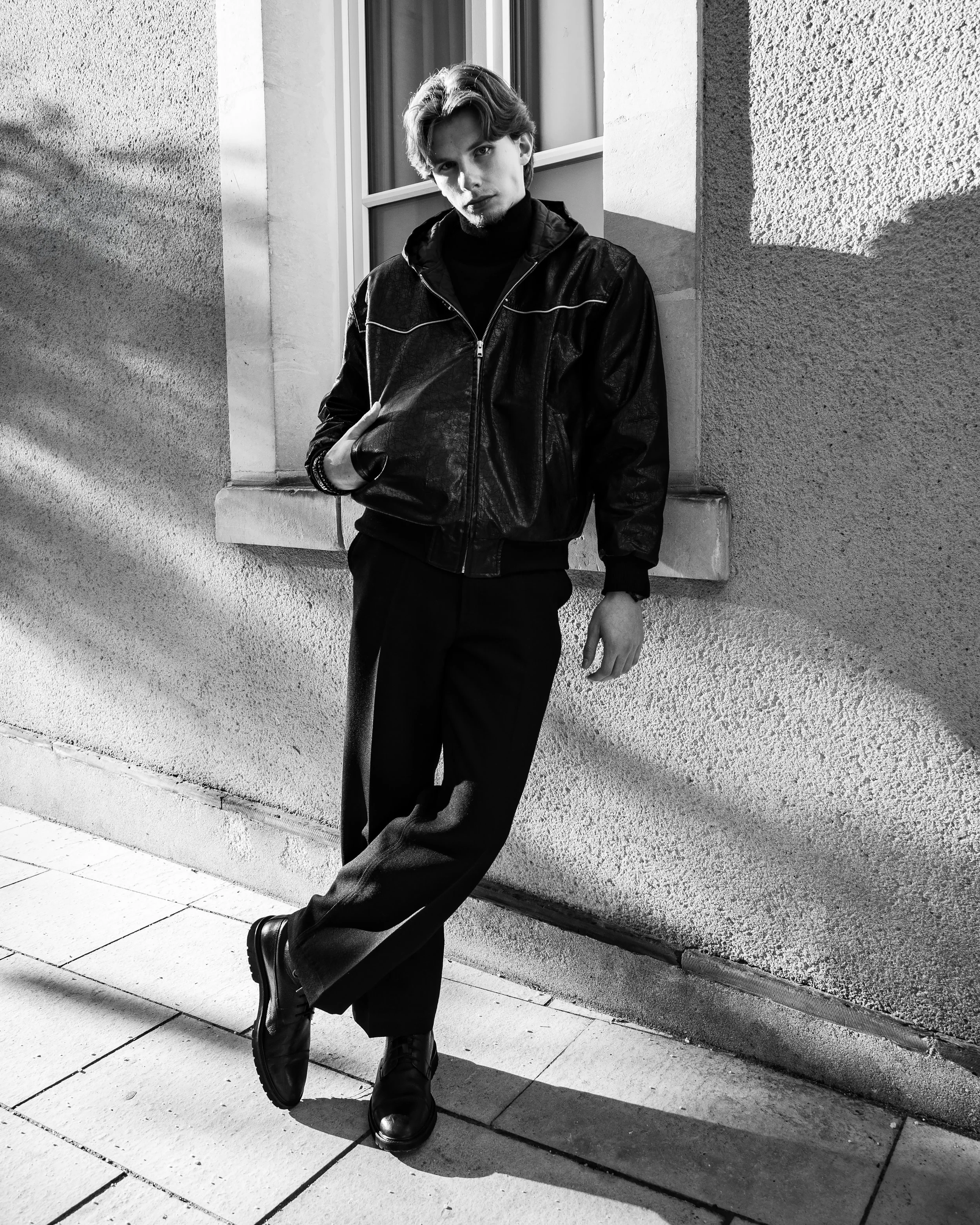 Un jeune homme debout contre un mur en extérieur, portant un blouson en cuir noir, une veste noire à col roulé et des bottes noires, dans une ambiance en noir et blanc.