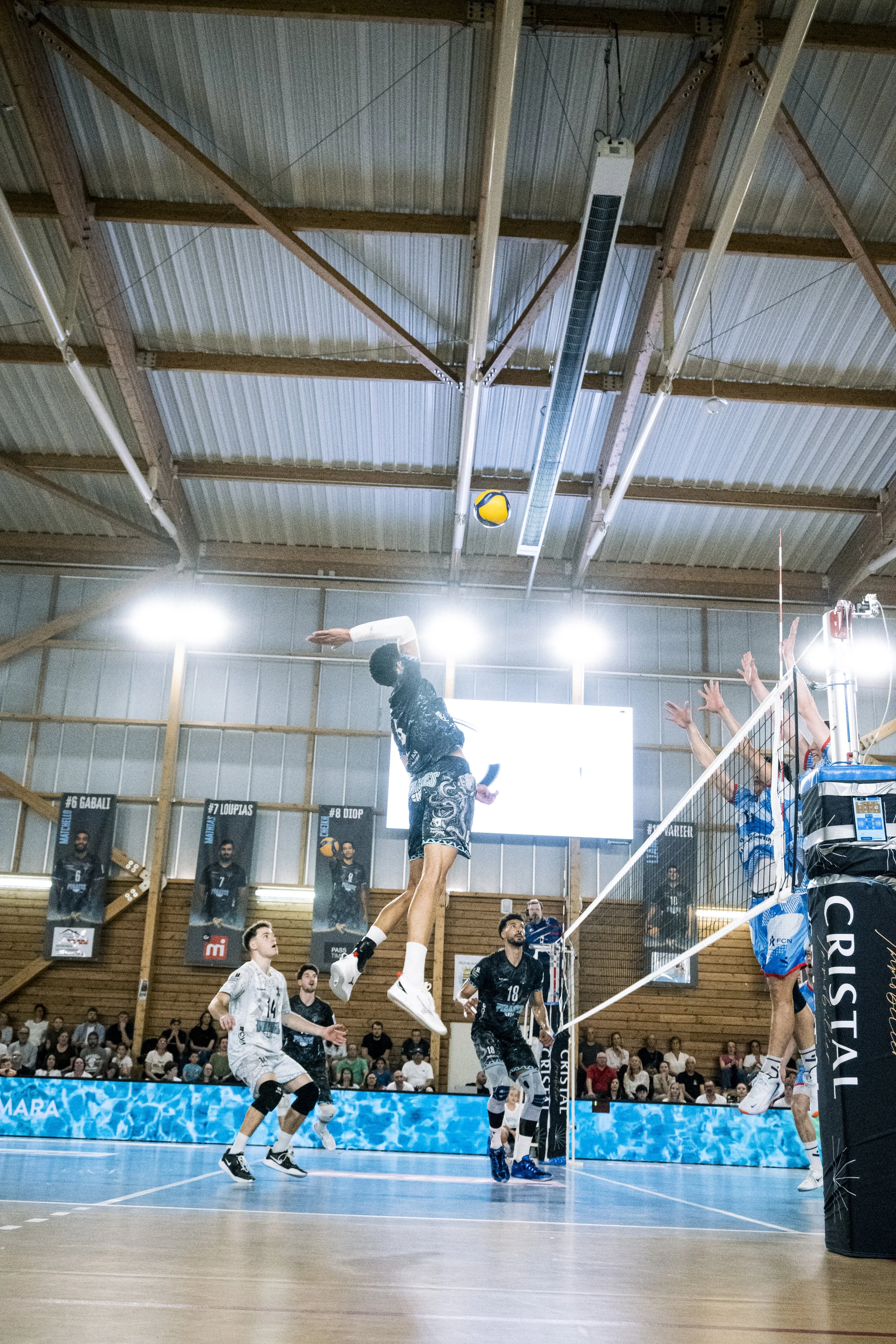 Une scène de volleyball en intérieur avec des joueurs en action, certains sautant pour bloquer ou attaquer, un public en arrière-plan et un panneau avec des noms de joueurs célébrés.