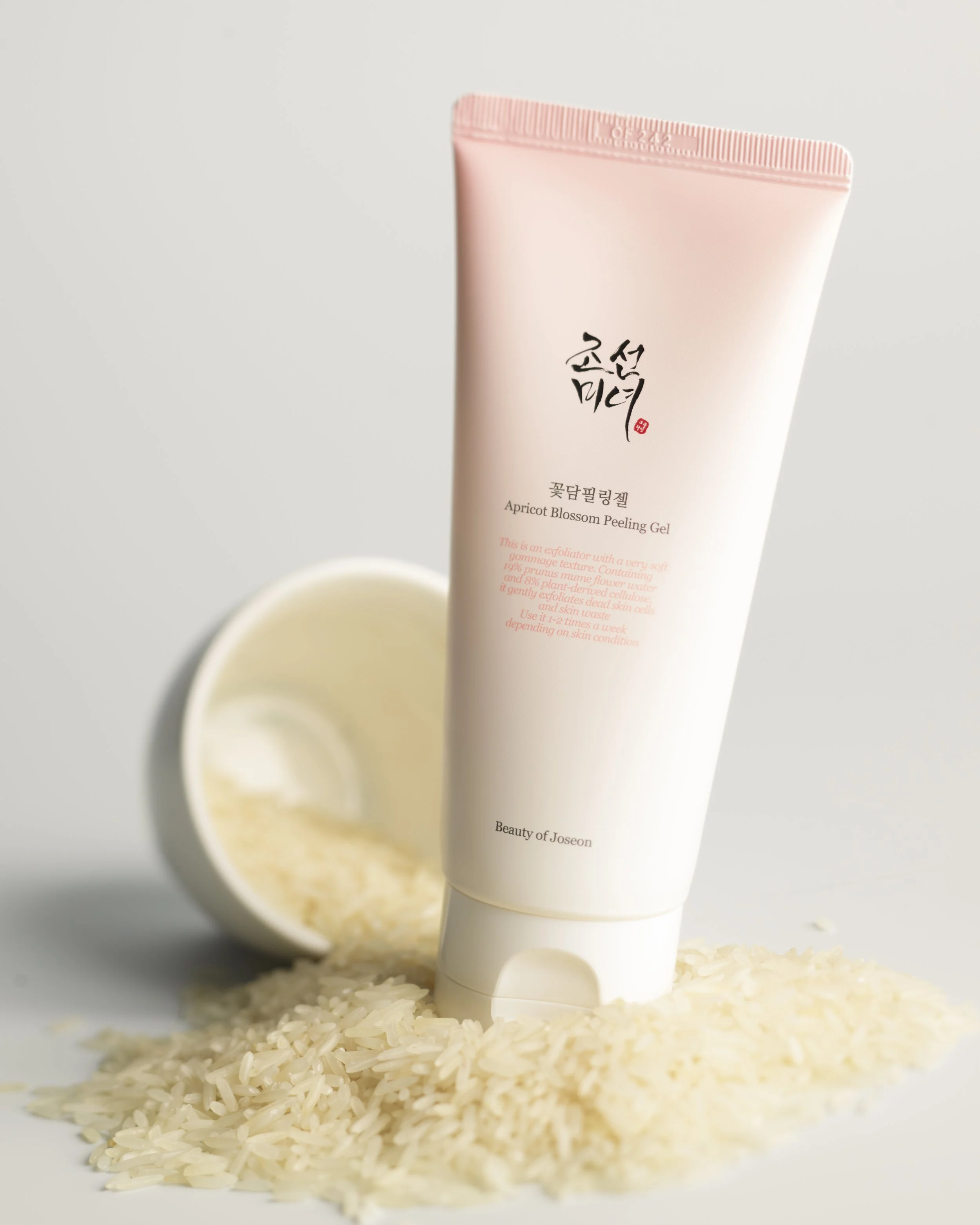 Une tube de gel peeling à la pêche en dessous d'un bol rempli de riz blanc, sur une surface blanche.