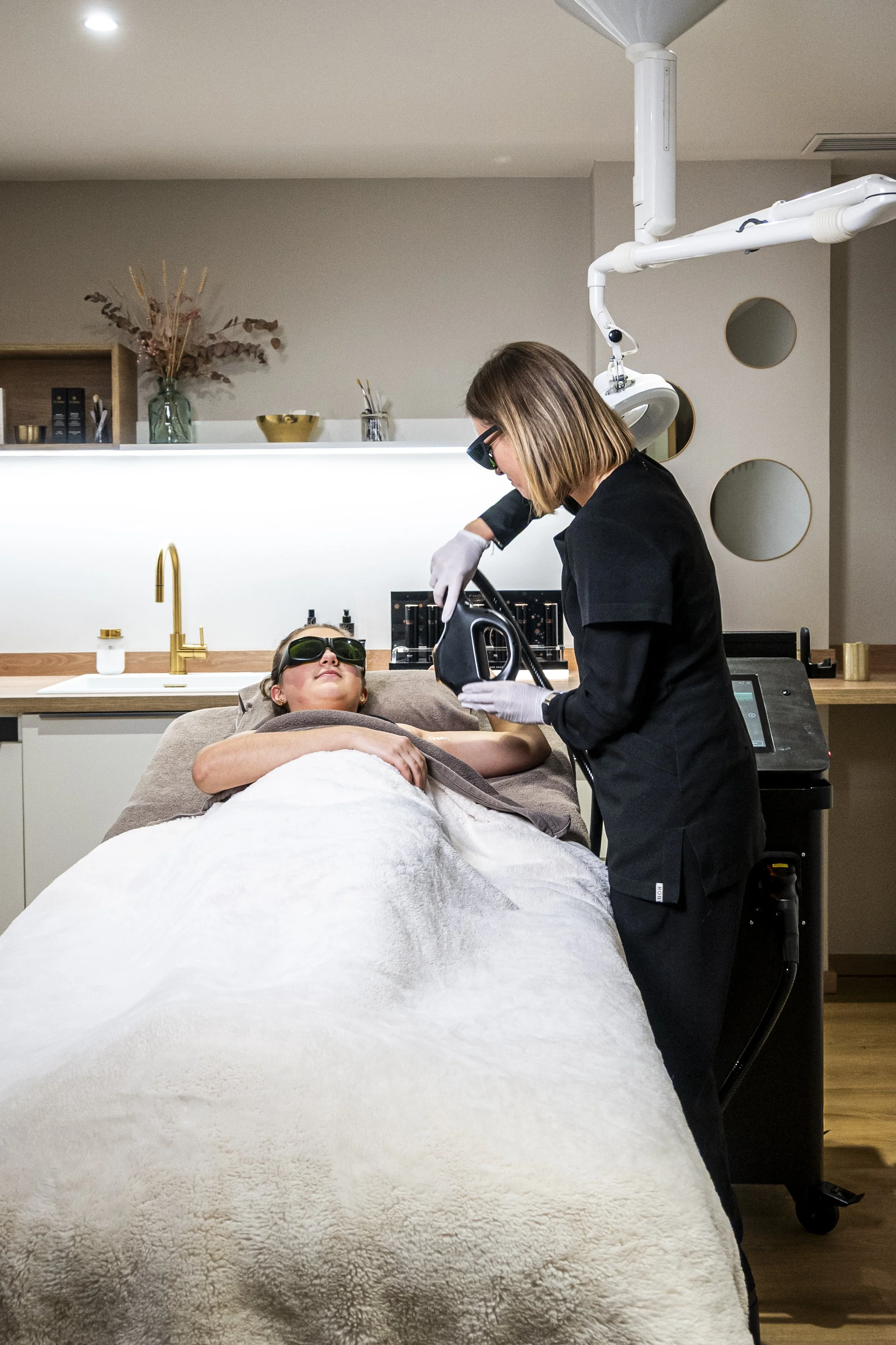 Une femme allongée sur une table de soin, portant des lunettes de protection, reçoit un traitement par une professionnelle dans un environnement moderne et épuré.