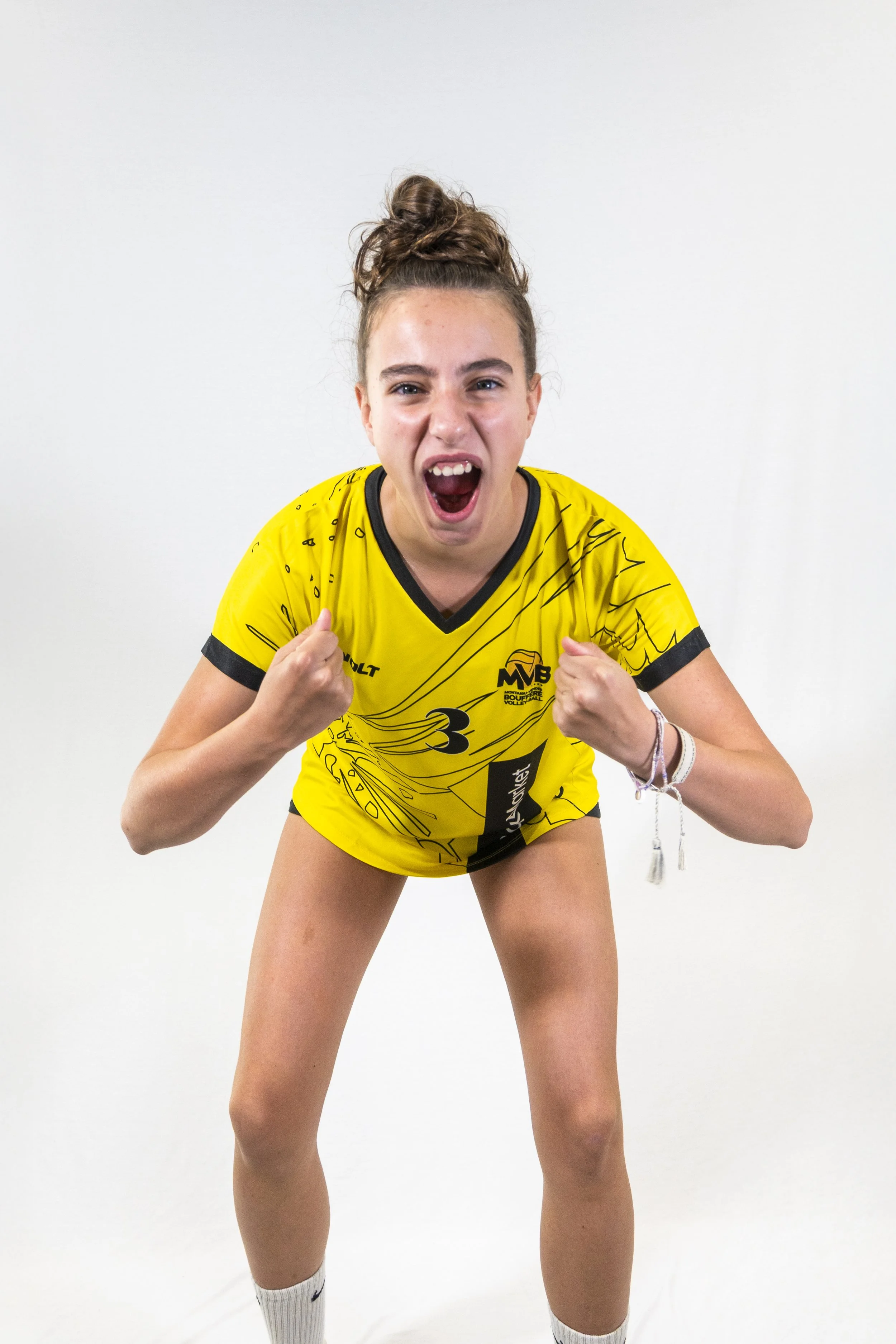 Jeune femme sportive en maillot jaune, les poings levés, criant d'enthousiasme, portant des chaussettes blanches et un bracelet, dans un fond blanc.