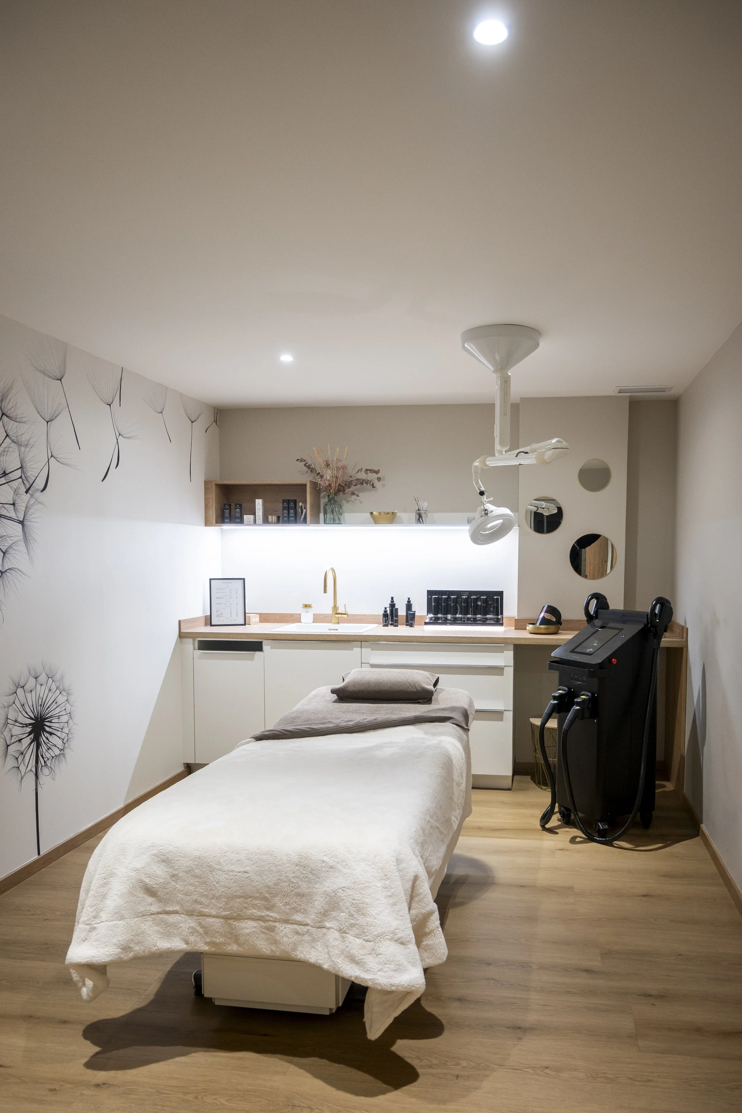 Salle de soins ou spa avec un lit d'examen recouvert d'une couverture blanche, appareils médicaux, décor moderne avec mur à motifs de pissenlits, étagère avec produits de soin, miroir rond et décoration florale.