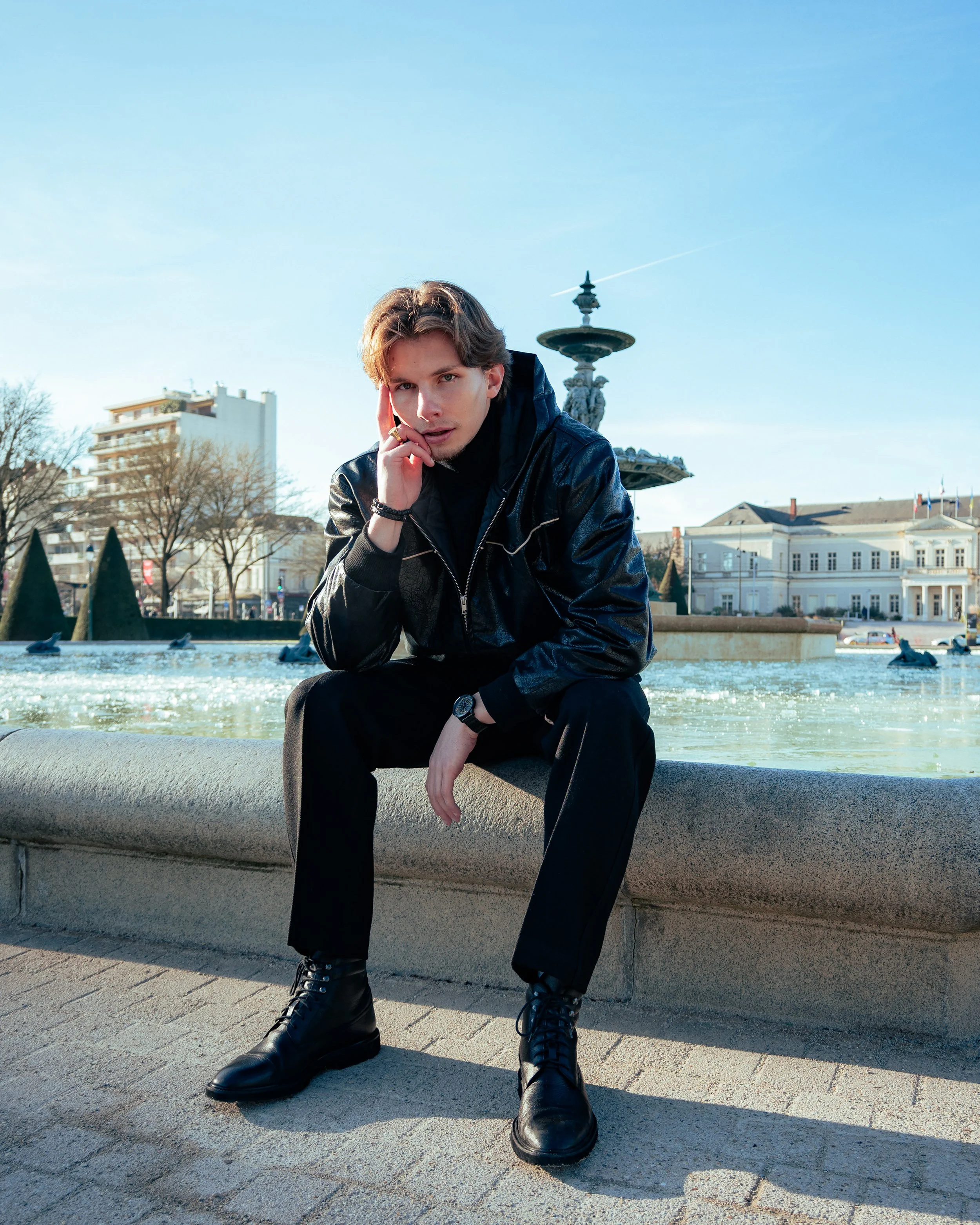 Jeune homme aux cheveux bruns, portant une veste en cuir noire et des bottes noires, assis près d'une fontaine dans un parc urbain en plein jour, avec des bâtiments en arrière-plan.