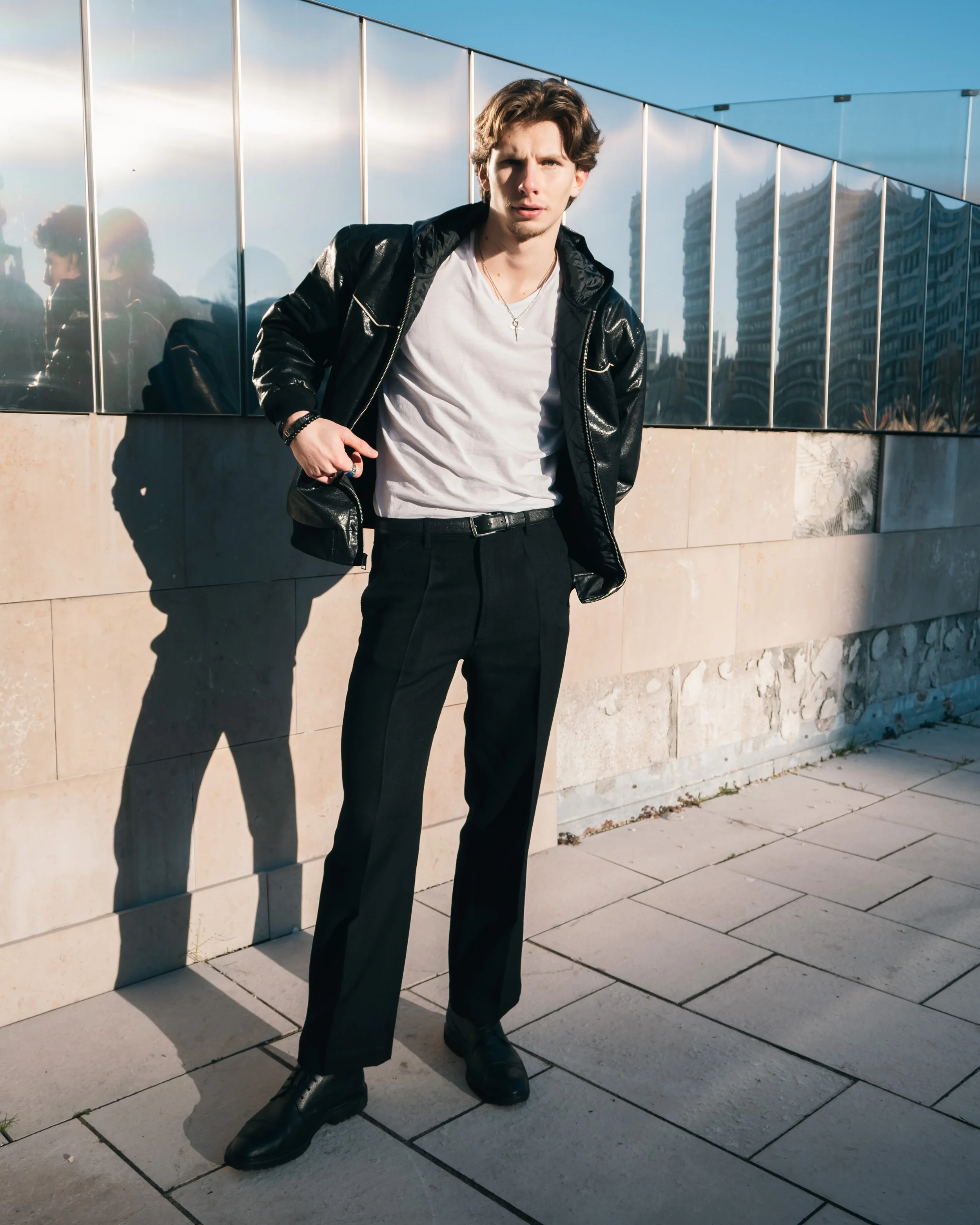 Jeune homme debout sur un trottoir avec un mur en verre derrière lui, portant une veste en cuir noire, un t-shirt blanc, un pantalon noir et des bottes, avec une expression perplexe. La reflection de l'homme est visible sur le mur en verre, et le paysage urbain se reflète dans le verre. Le ciel est clair et ensoleillé.