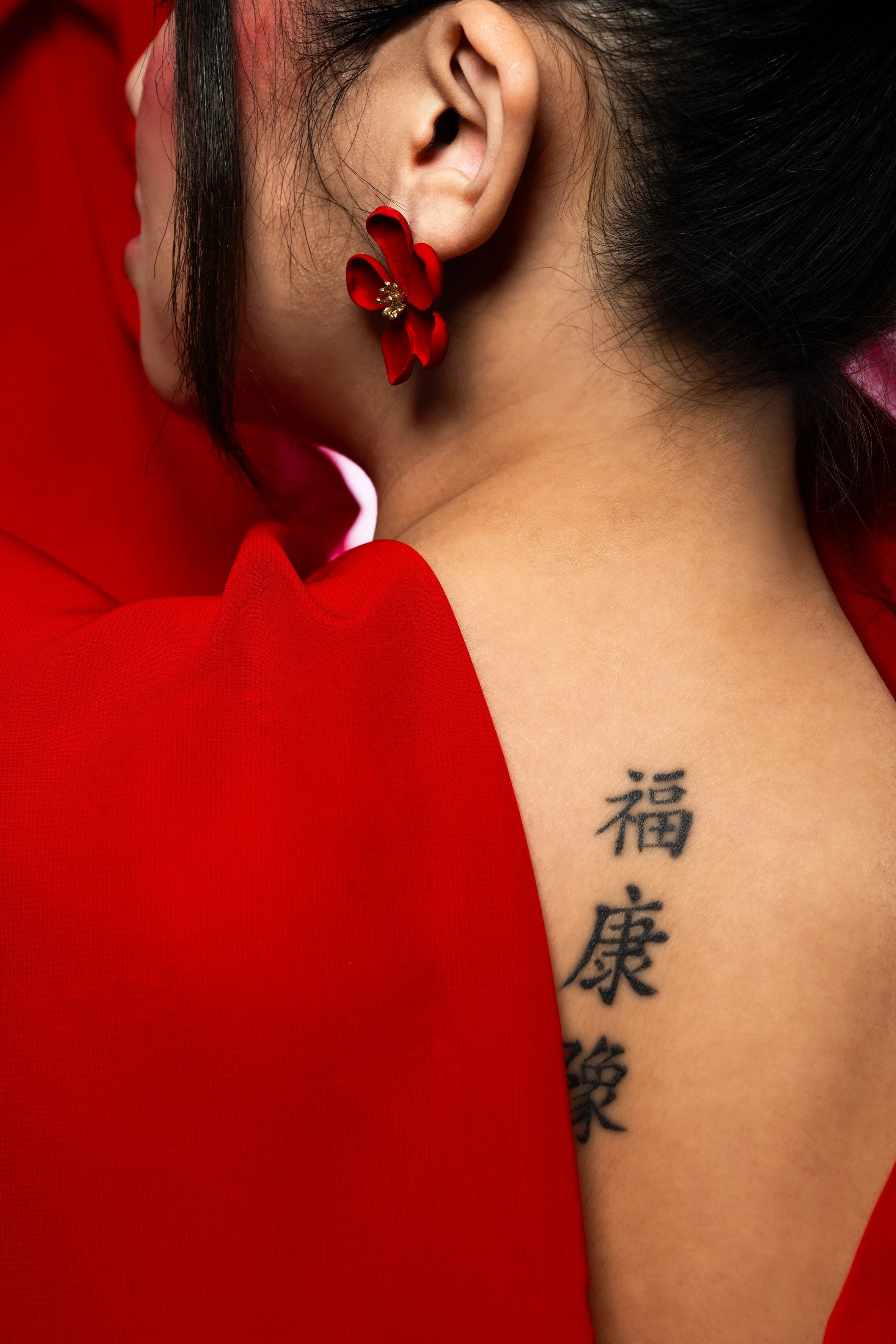 Close-up de l'oreille d'une femme portant une boucle d'oreille en forme de fleur rouge avec des détails dorés. La femme porte une robe rouge et a une tatouage chinois en noir sur le bras.