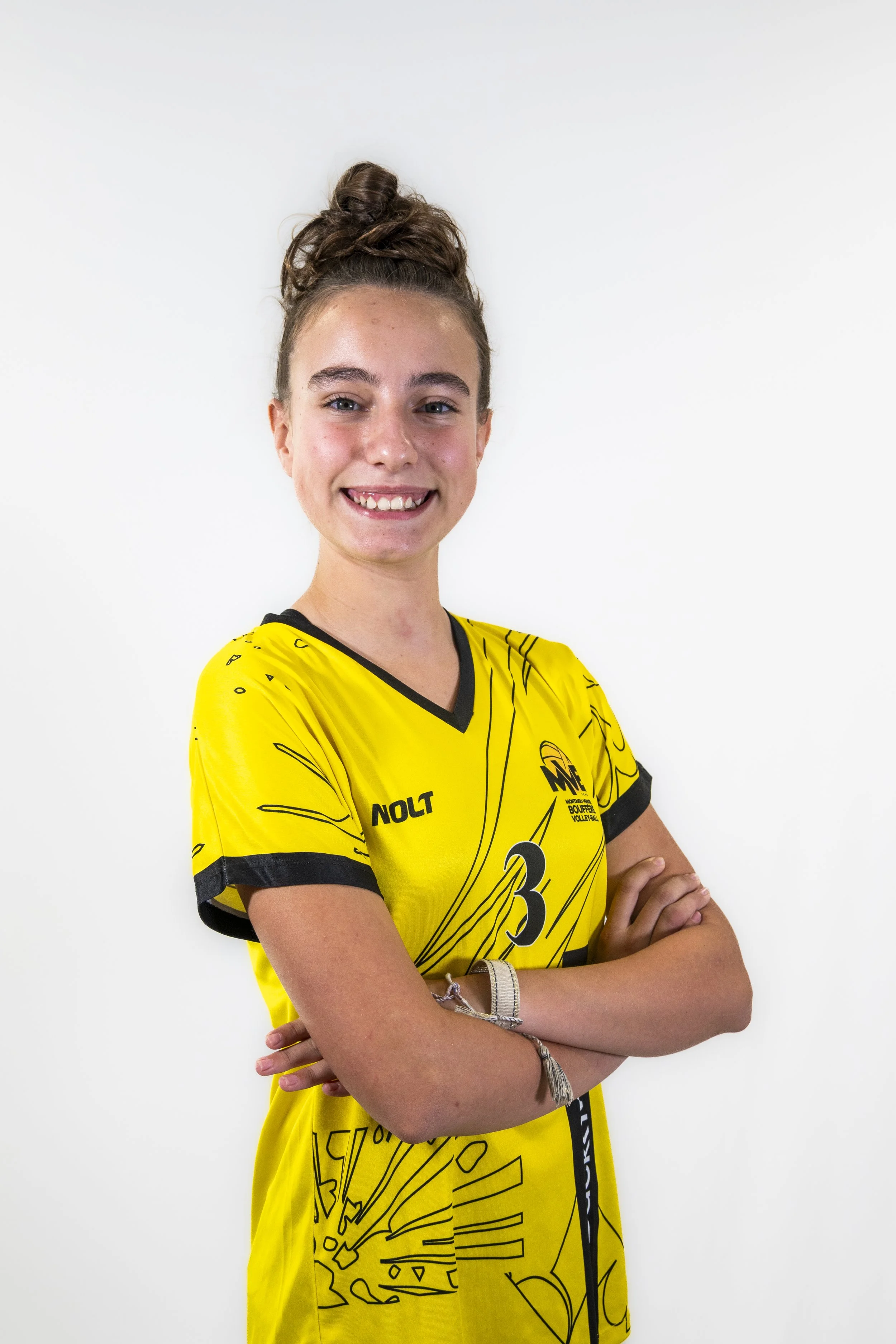 Une jeune femme souriante, portant un maillot de volleyball jaune avec des motifs noirs, les bras croisés, sur un fond blanc.