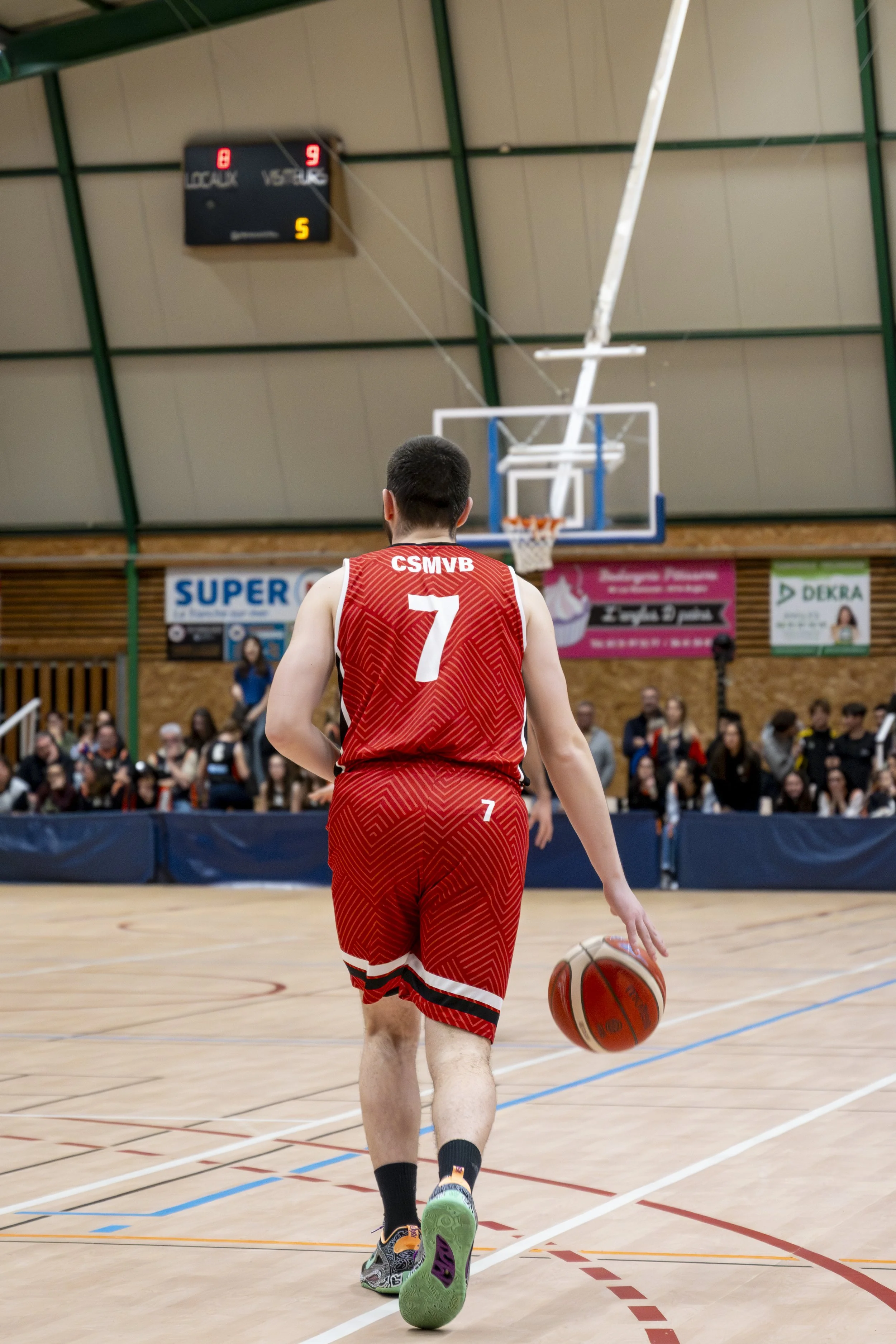 Un joueur de basketball vêtu d'un maillot rouge portant le numéro 7, dribble un ballon sur un terrain intérieur lors d'un match, avec des spectateurs en arrière-plan.