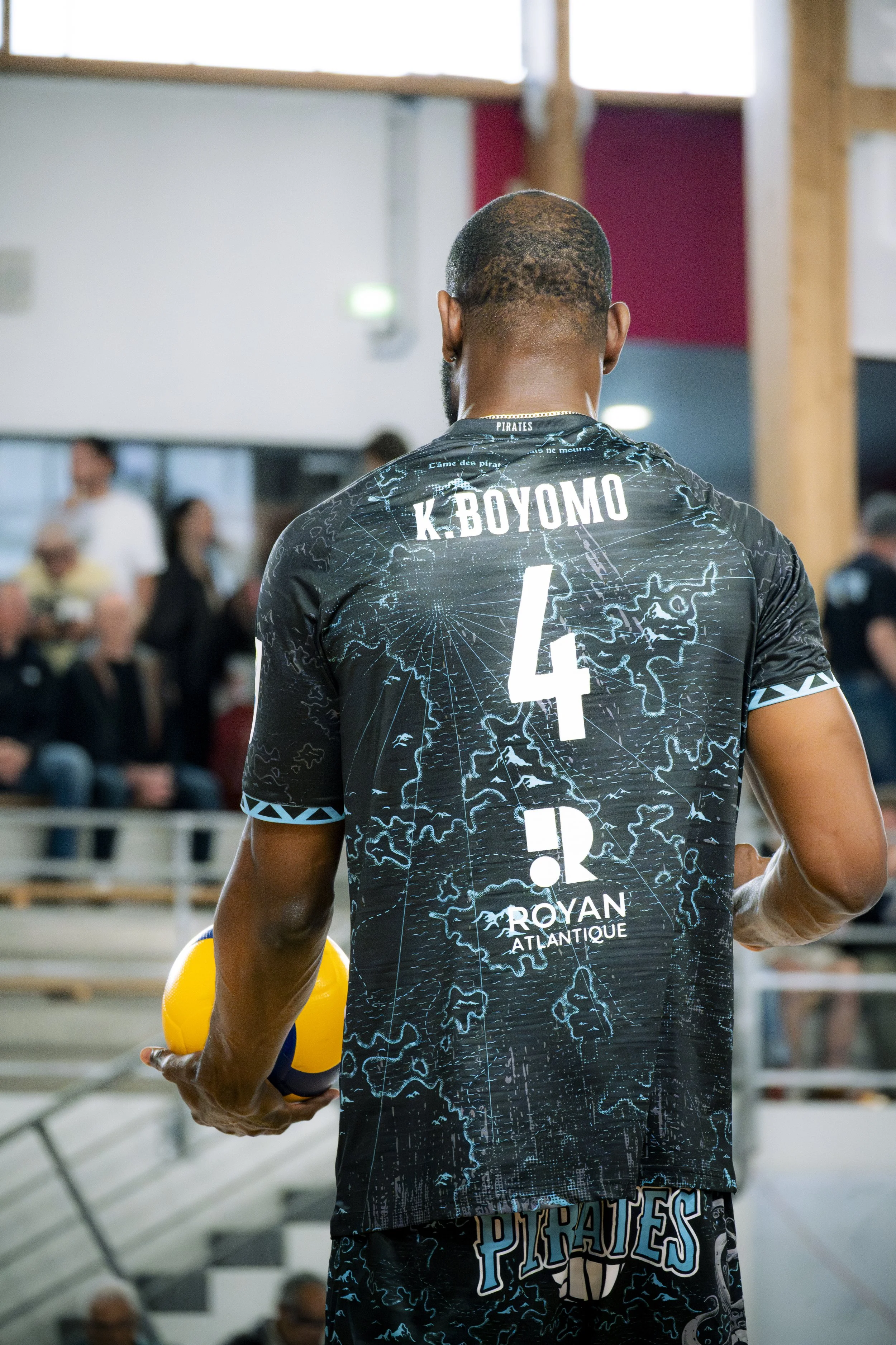Un joueur de volleyball vu de derrière, portant un maillot noir avec des inscriptions et un nom, tenant un ballon yellow et blue, dans un gymnase, avec des spectateurs en arrière-plan.