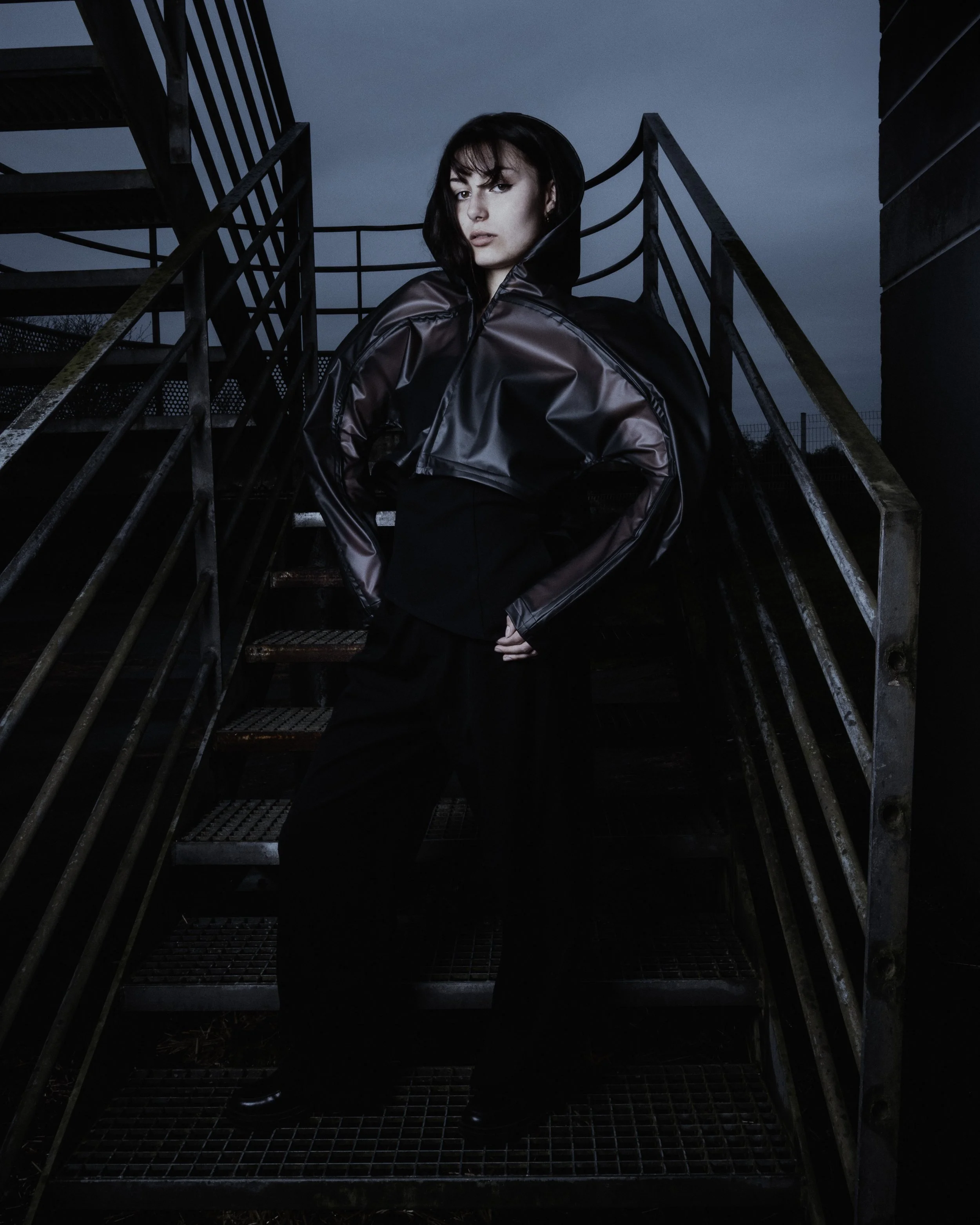 Une femme portant un manteau en plastique sombre, debout au sommet d'une structure métallique d'escalier en extérieur, avec un ciel sombre en arrière-plan.