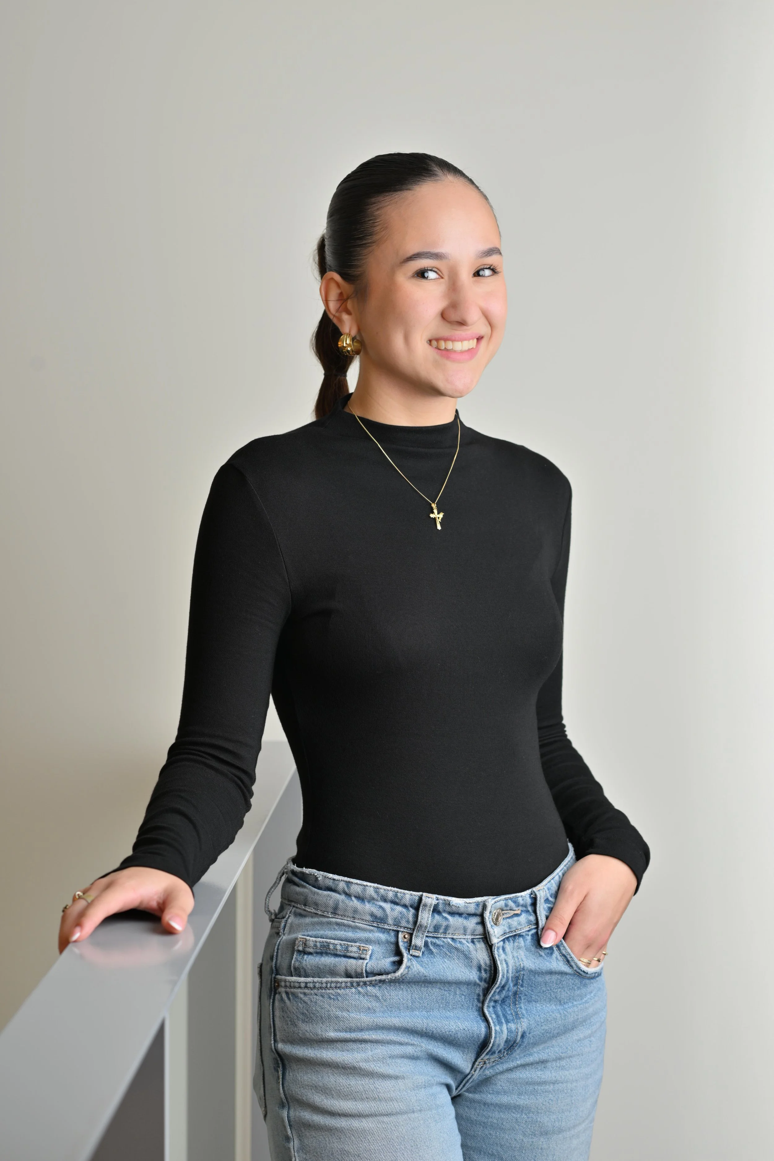 Jeune femme souriante portant un t-shirt noir, des bijoux dorés, et un jean, posant contre un mur blanc.