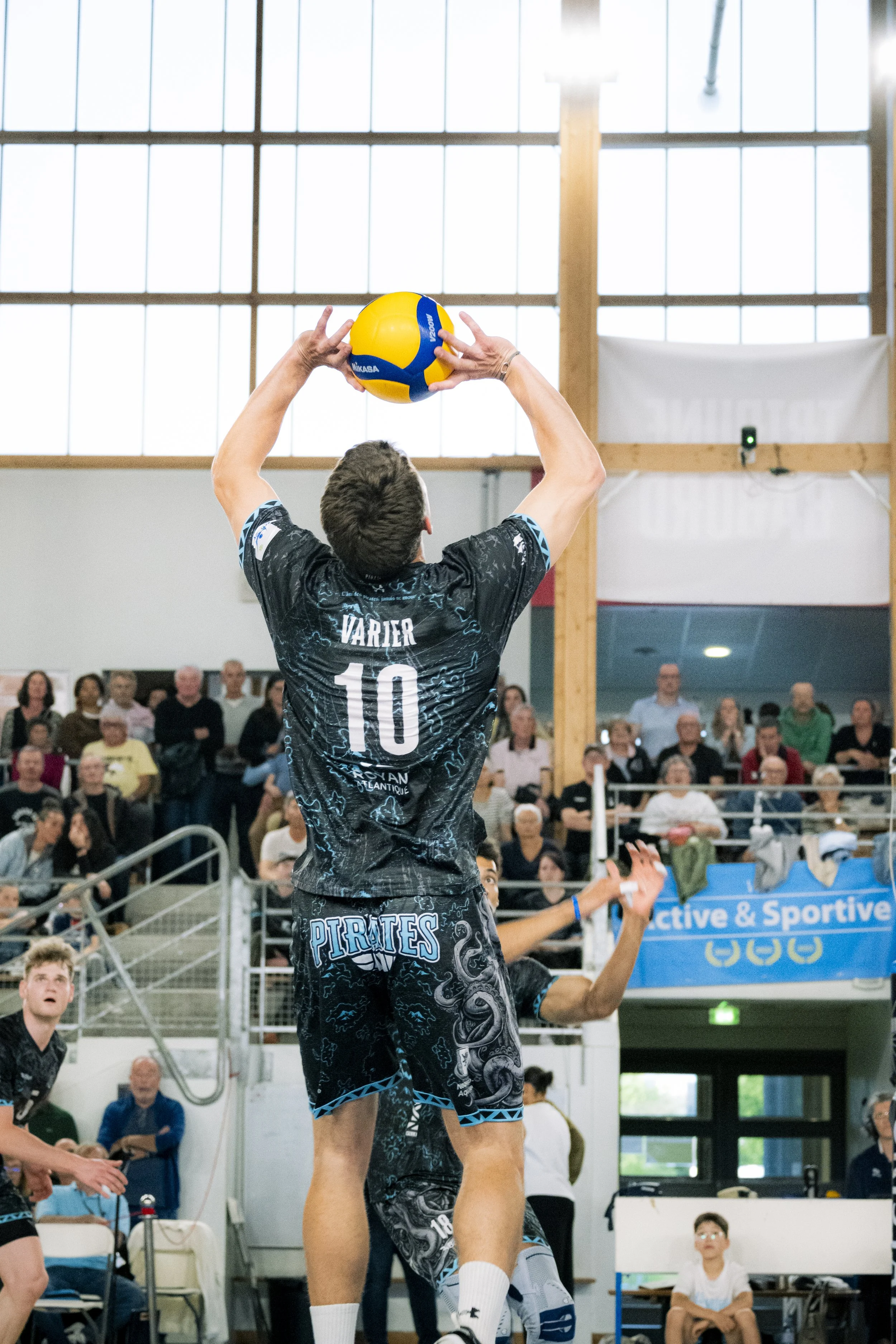 Un joueur de volleyball en action lors d'un match en intérieur, portant un maillot noir avec le nom "Vartier" et le numéro 10. Il attrape un ballon de volleyball jaune et bleu. Le public regarde le match dans les gradins.