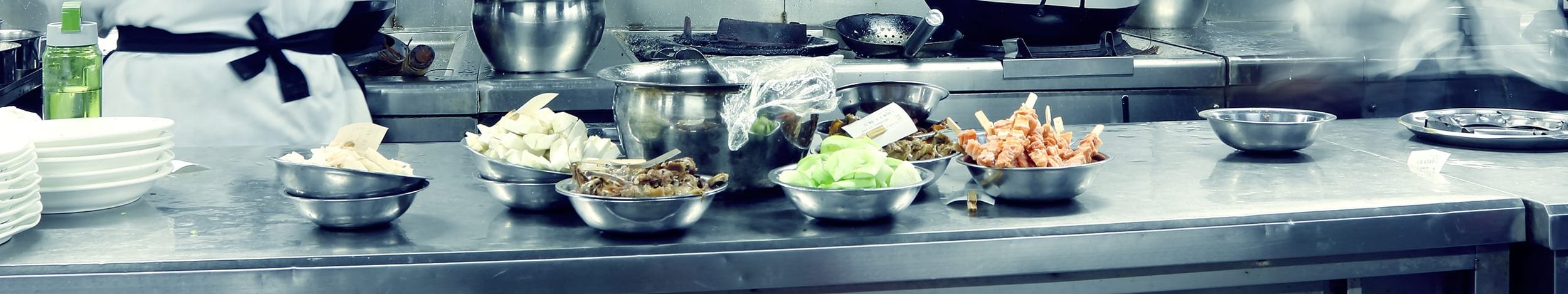 Kitchen counter with bowls of chopped vegetables, meat, and working chef in the background preparing food.