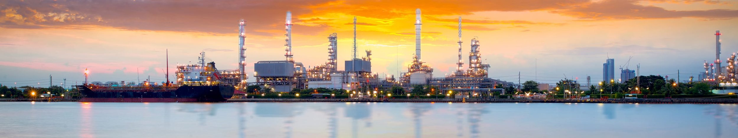 Industrial oil refinery with multiple tall pipes and a docked ship, reflecting on a calm body of water during sunset.