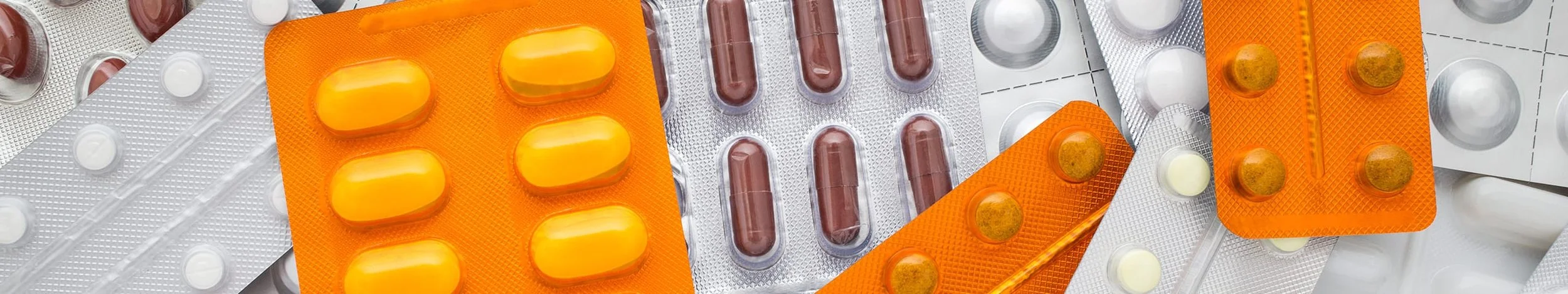 Various blister packs of pills and capsules in different shapes, sizes, and colors, including yellow, brown, and white, scattered randomly.