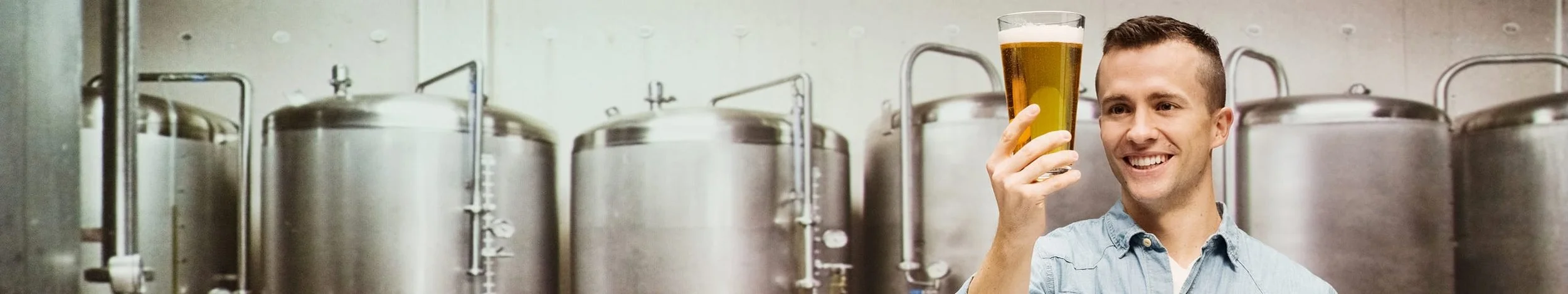 A man in a light blue shirt holding a glass of beer in front of industrial brewing tankers.
