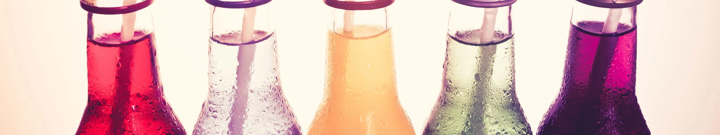 Five glass soda bottles filled with colorful liquids, with straws inside, arranged in a row.