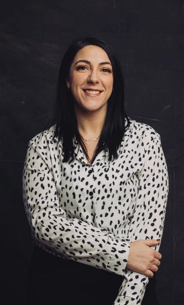 Woman with dark hair wearing a white polka dot blouse, smiling with arms crossed against a dark background.