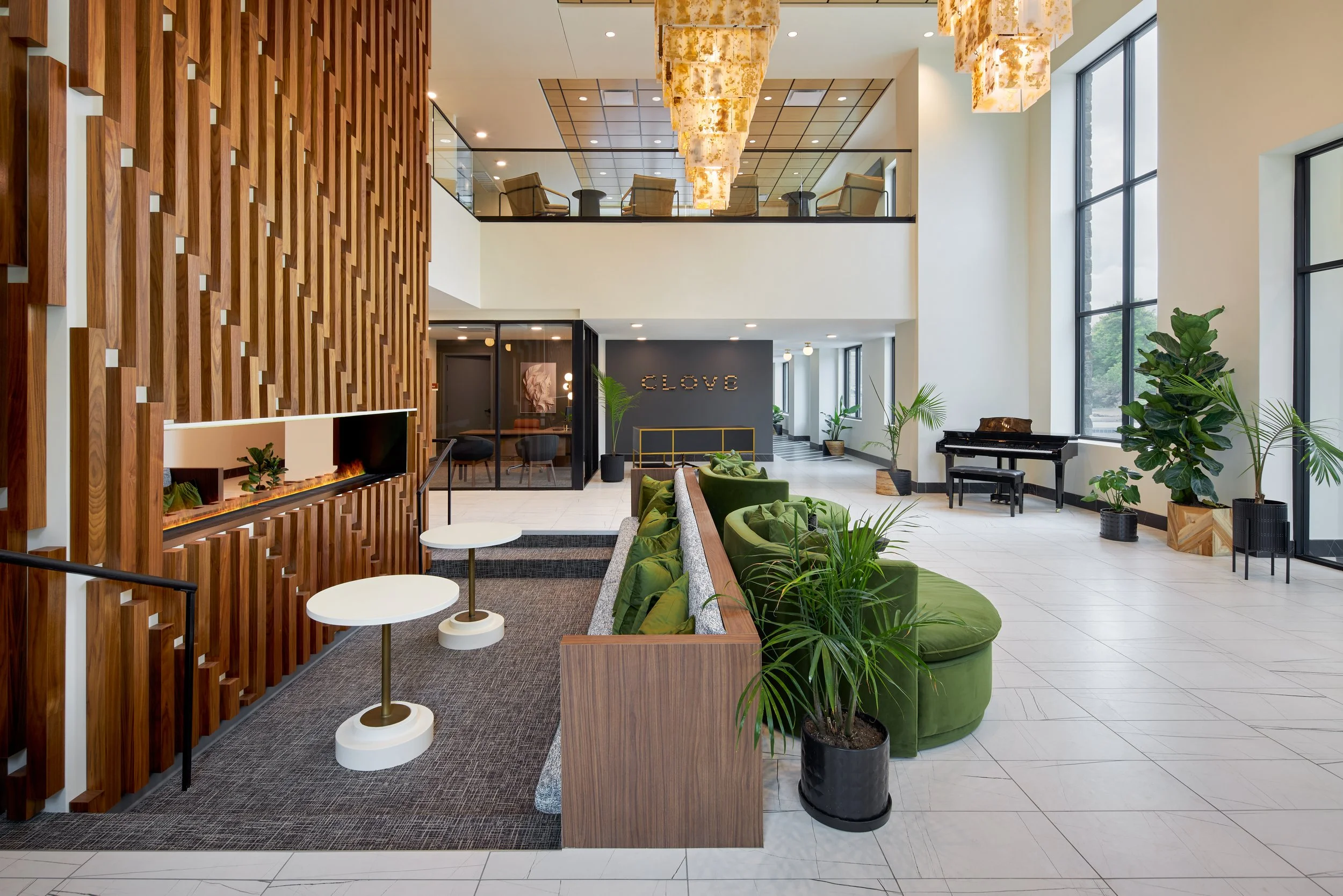 Modern lobby with wooden wall accents, green seating area, round white tables, large windows, hanging lights, and indoor plants; grand piano in the background.
