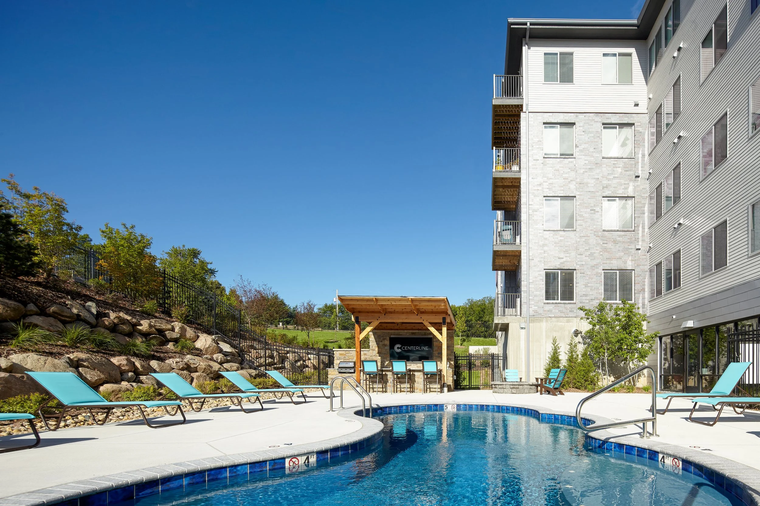 Outdoor pool area at an apartment complex with lounge chairs and a shaded seating area, adjacent to a modern building.