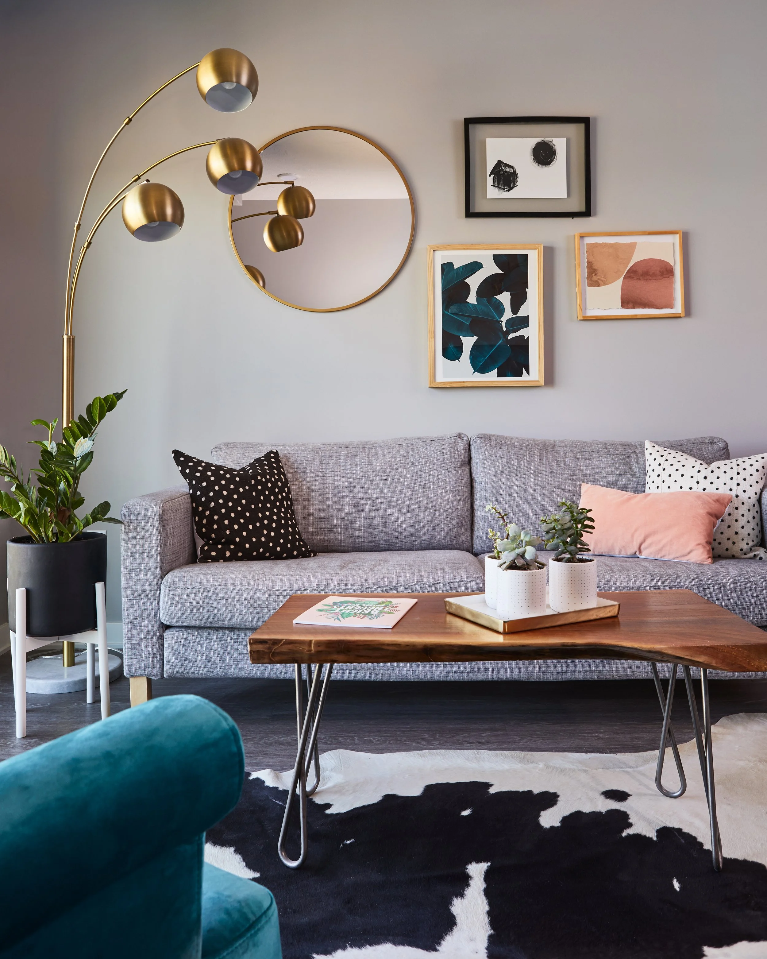 Modern living room with gray sofa, throw pillows, wooden coffee table, indoor plants, gold floor lamp, abstract wall art, and circular mirror on the wall.