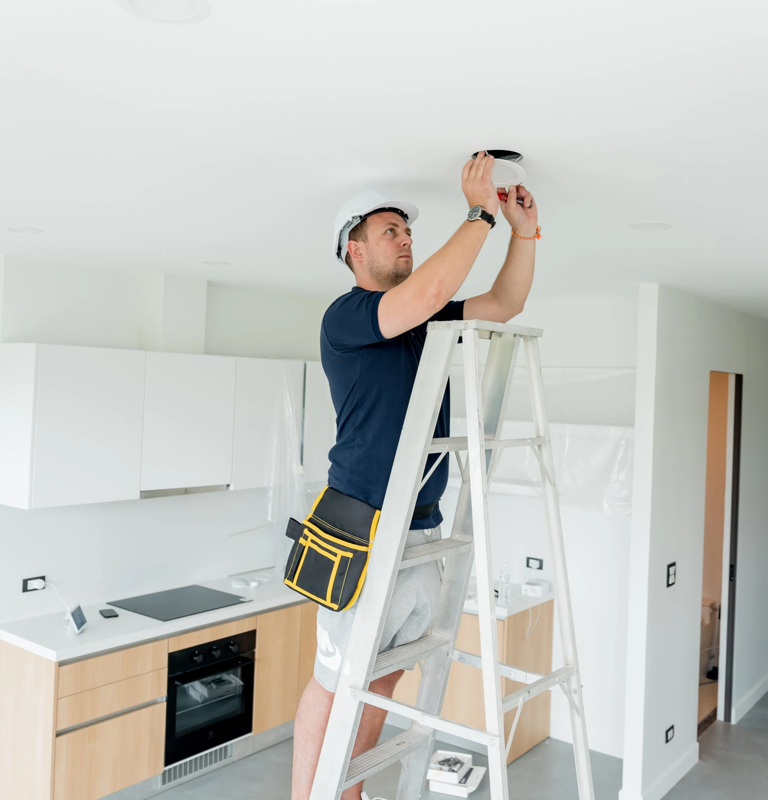 A man on a ladder installing a smoke detector on the ceiling in a modern kitchen, wearing a hard hat and tool belt.