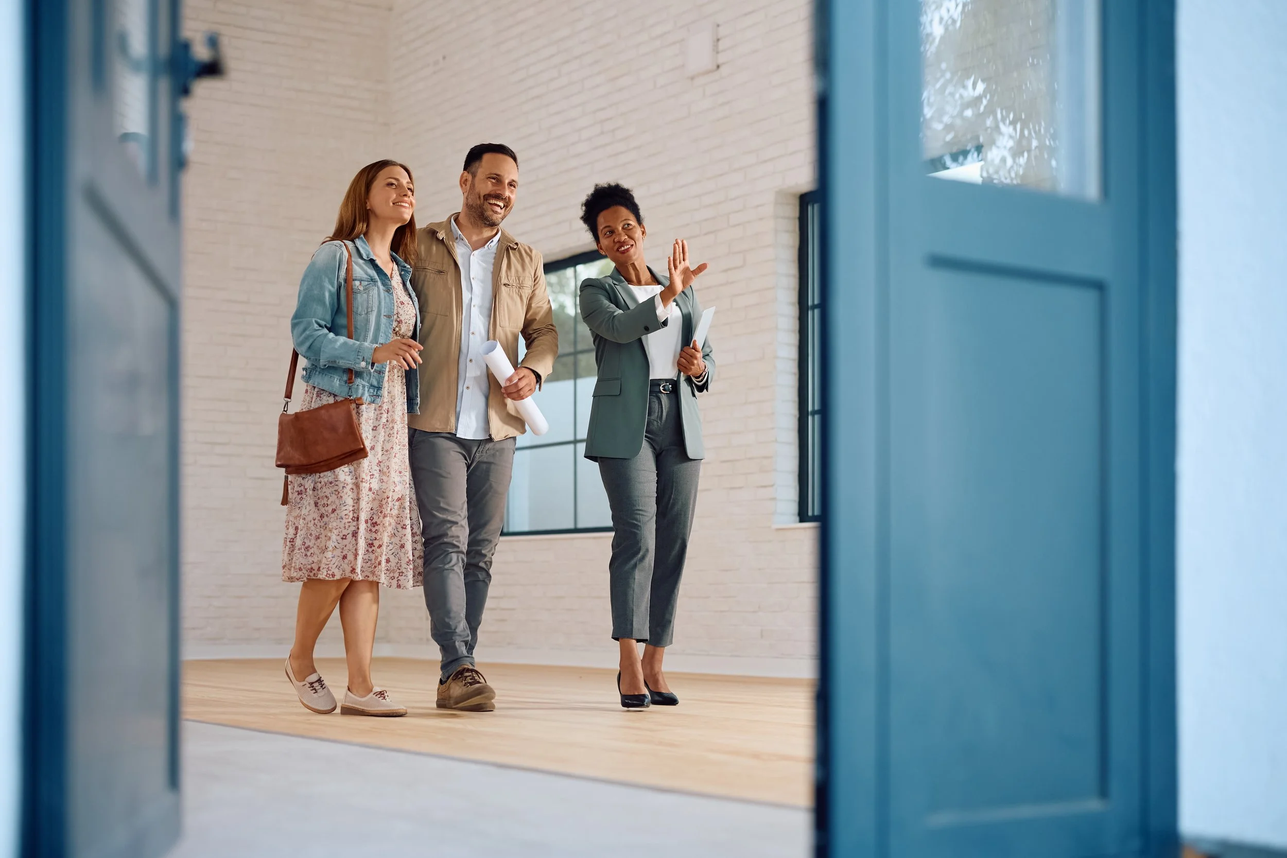 A couple touring a home with a real estate agent, standing in a bright room with wooden floors and white brick walls.
