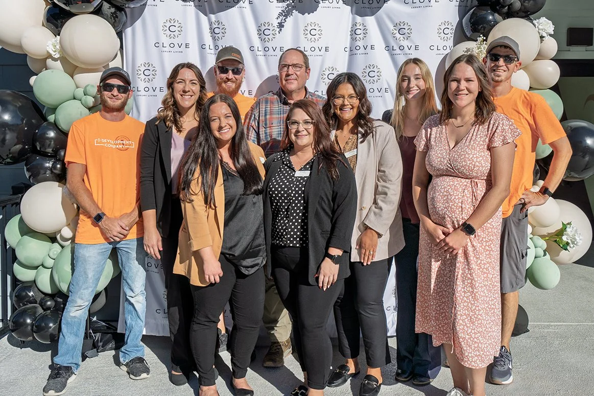 A group of ten people posing in front of a "CLOVE Luxury Living" backdrop with balloon decorations. They are standing outdoors in a sunny environment, dressed in casual and business attire.