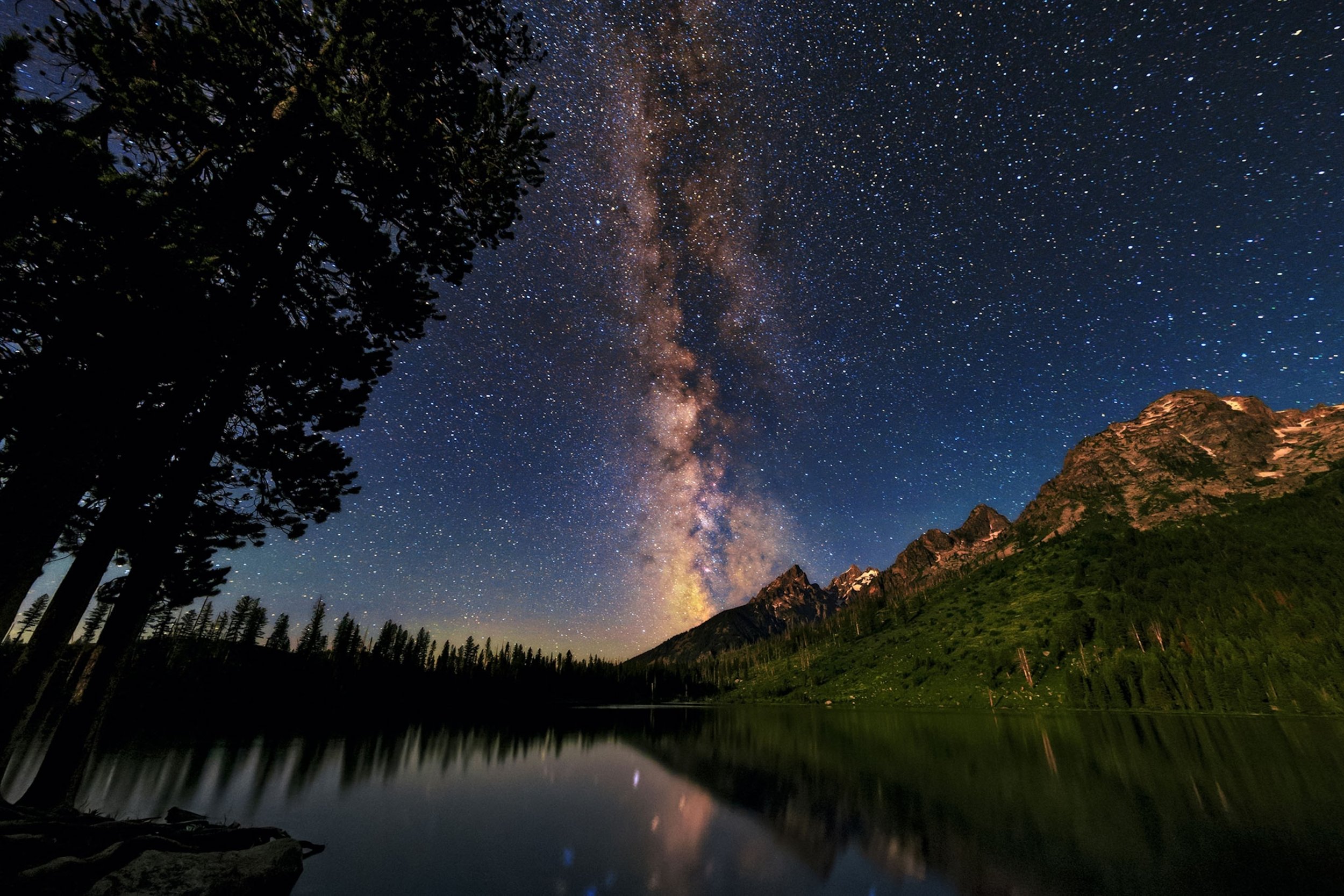 Starry night sky with the Milky Way galaxy visible above mountains and a reflective lake with trees in the foreground.