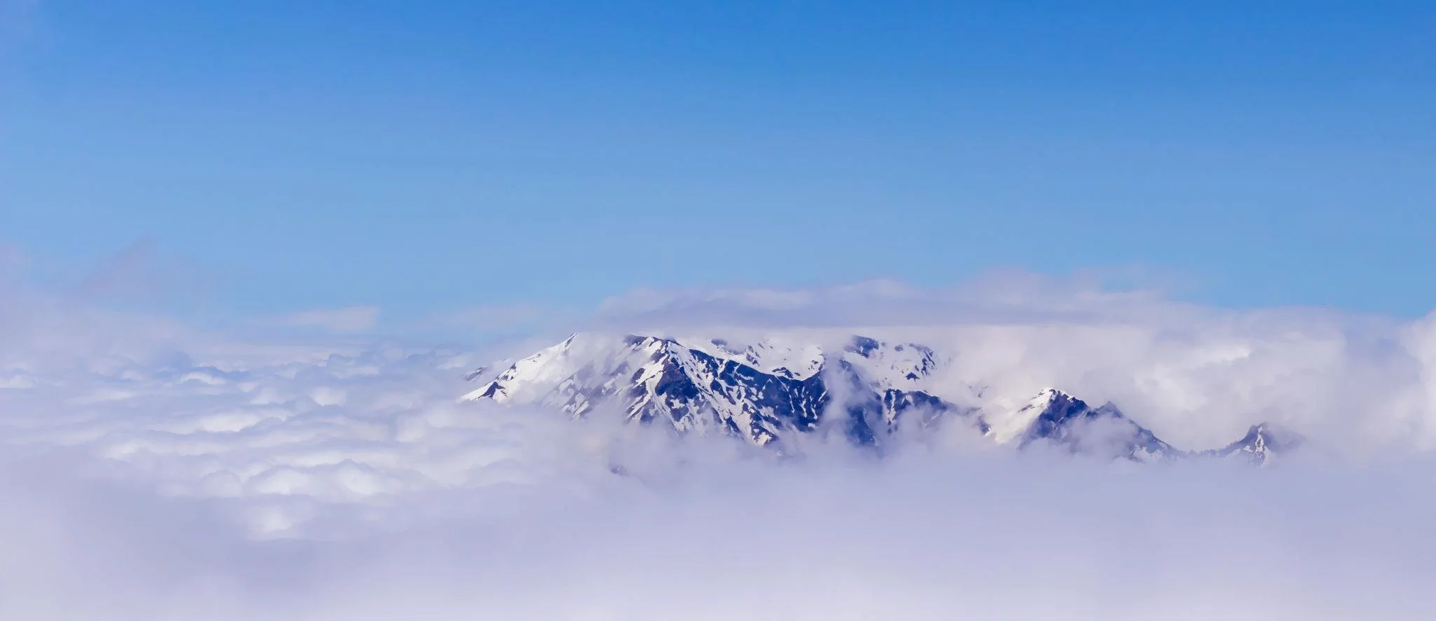 Snow-capped mountain peak surrounded by clouds under a clear blue sky.