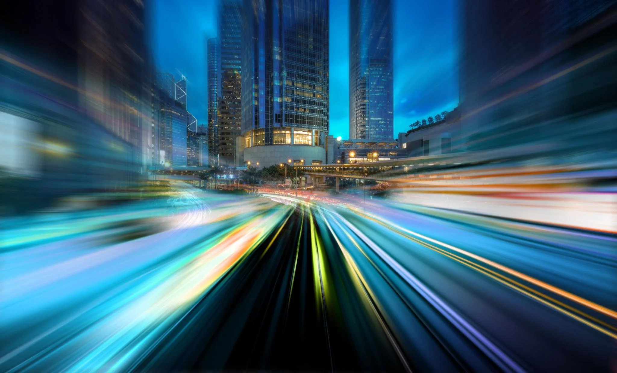 Night cityscape with tall glass skyscrapers and colorful light trails from moving vehicles.