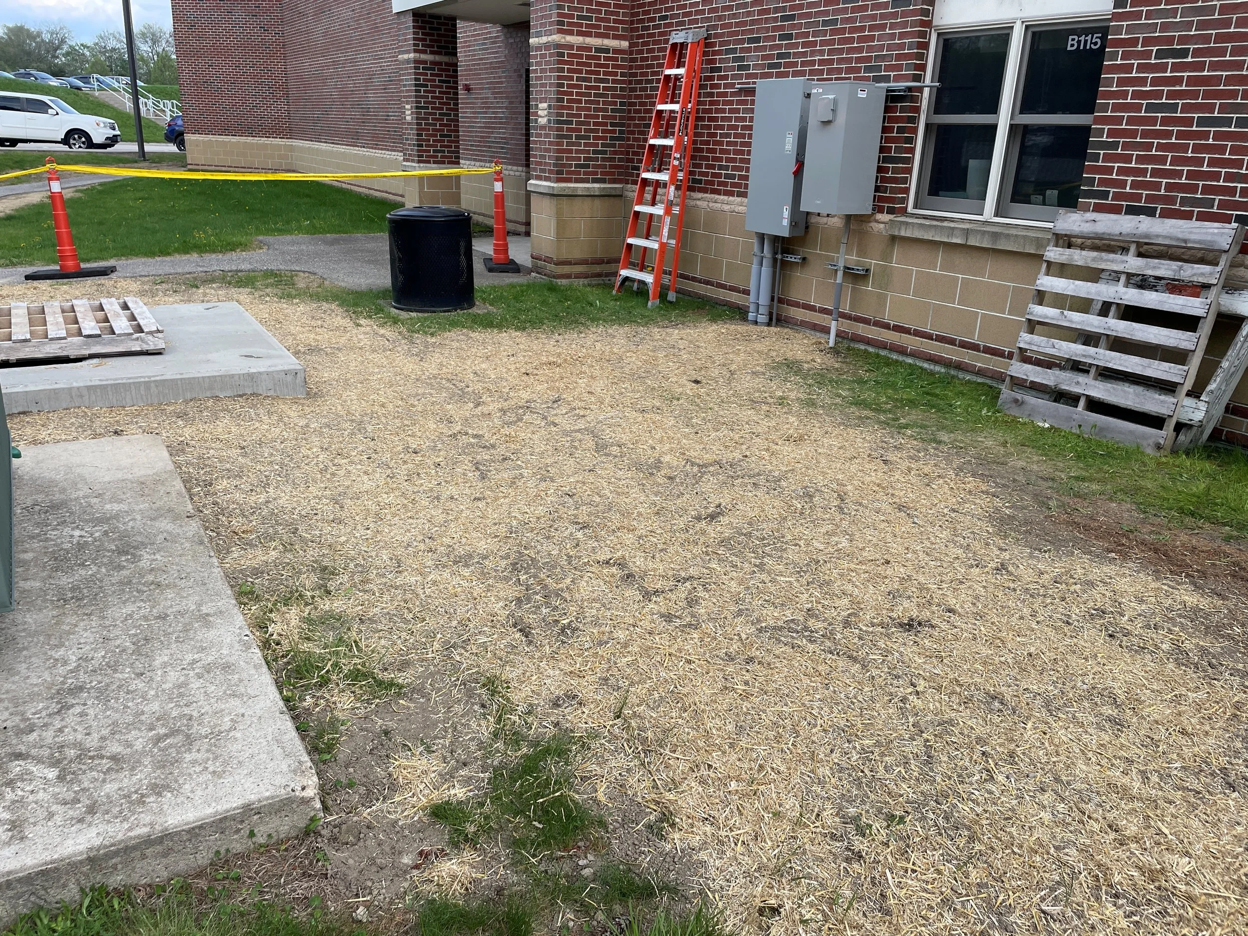 Construction area outside of a brick building with orange cones, yellow caution tape, a ladder, and construction materials, including a pallet and utility box.