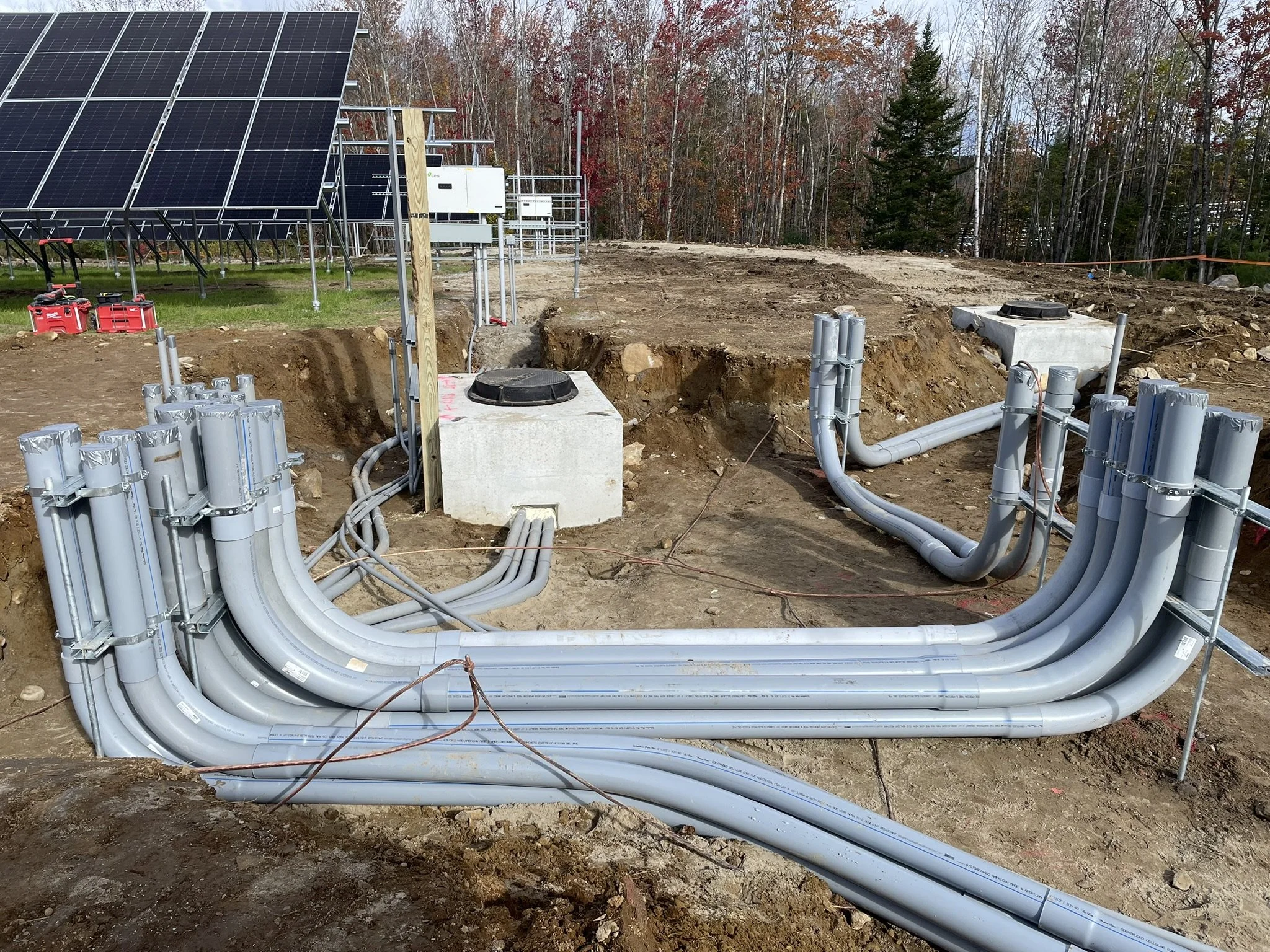 Installation of electrical wiring and equipment for solar panel system outdoors, with solar panels visible in the background, trees, and uneven ground.