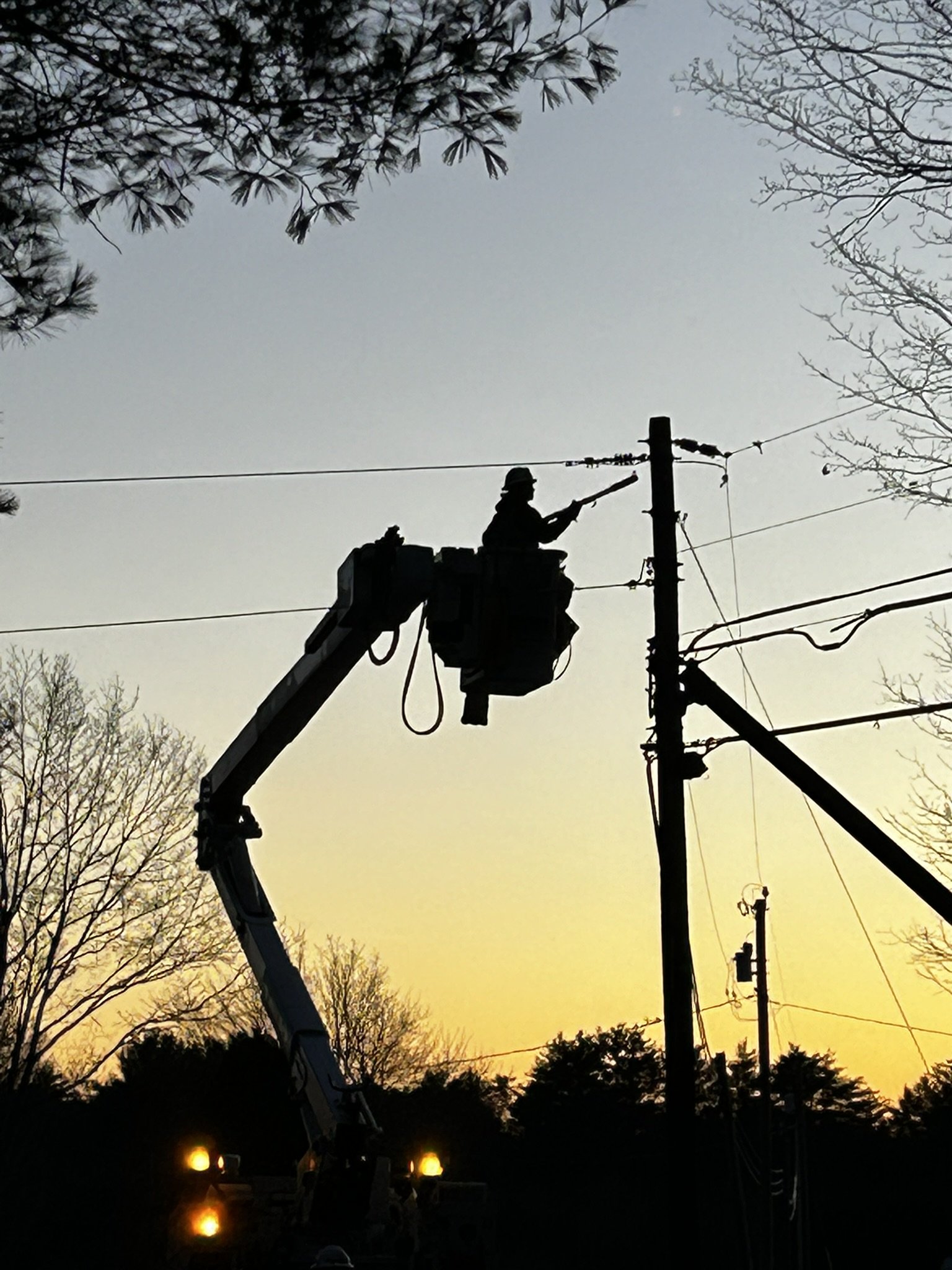 A silhouette of a utility worker on a lift bucket working on power lines during sunset, with trees and a clear sky in the background.