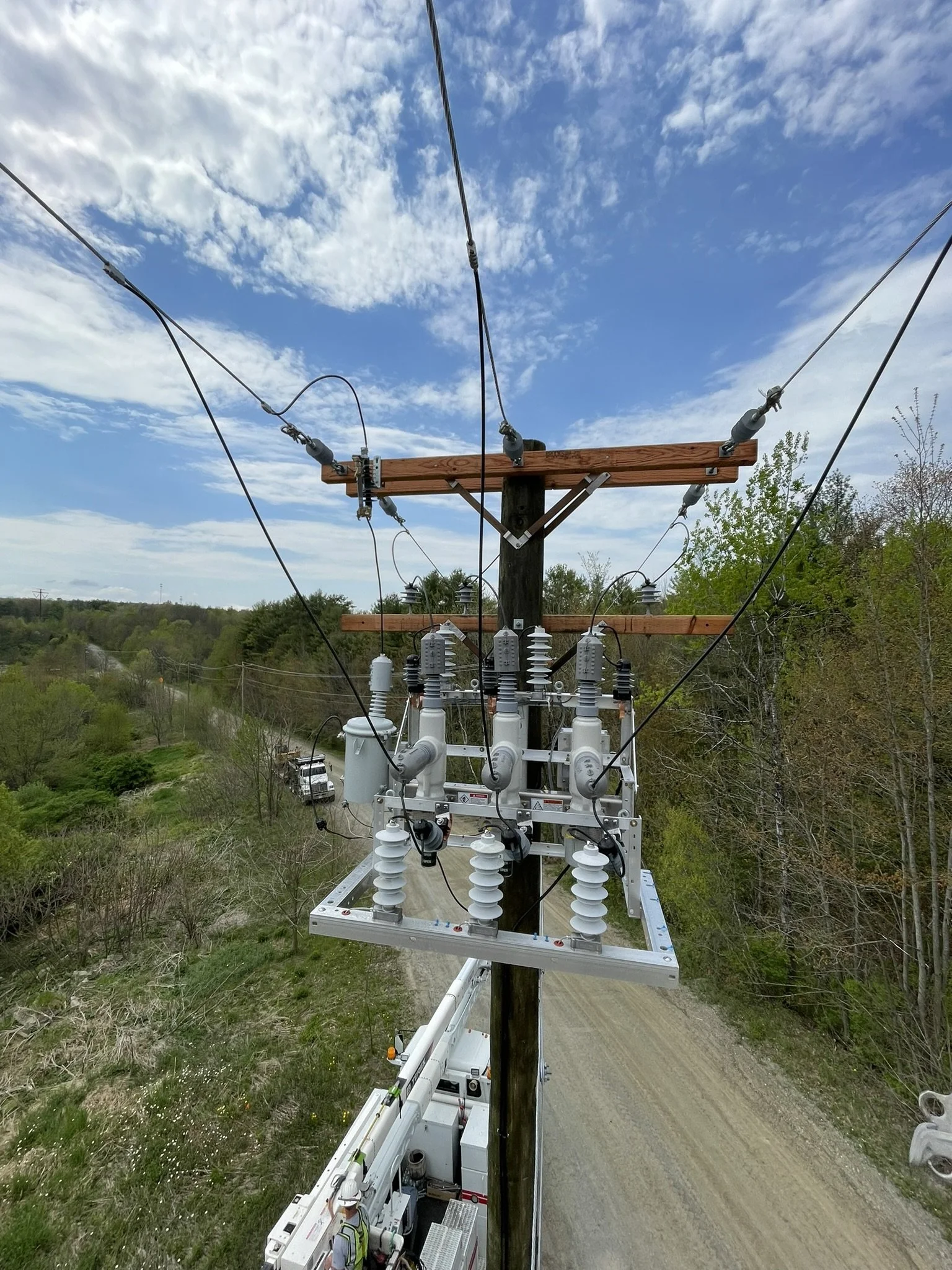 A utility worker in a safety vest and helmet working on a utility pole with electrical equipment, wires, and insulators, near a rural road under a partly cloudy sky with trees in the background.