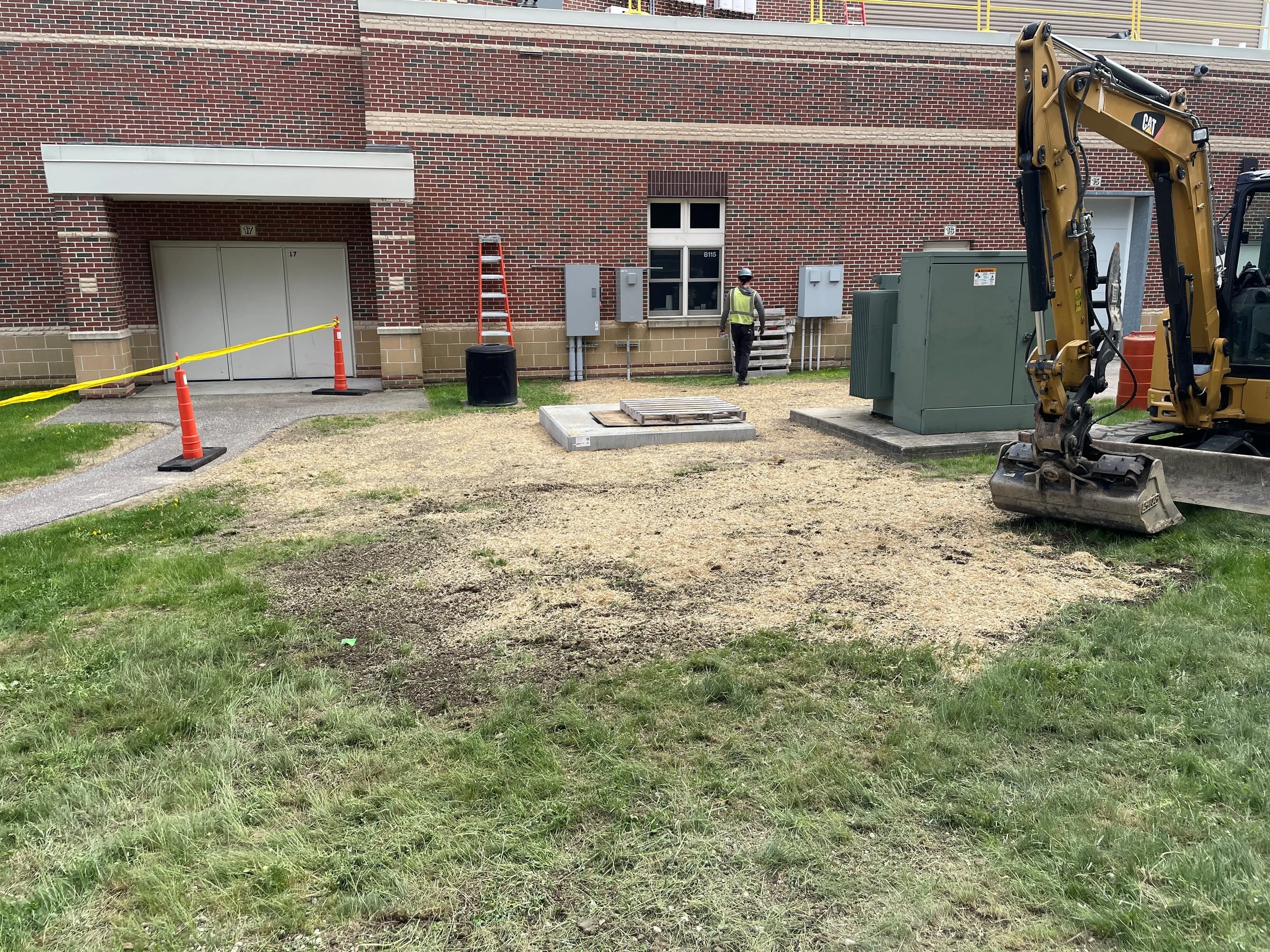 Construction worker wearing a safety vest and helmet walking near construction equipment and warning cones outside a brick building.