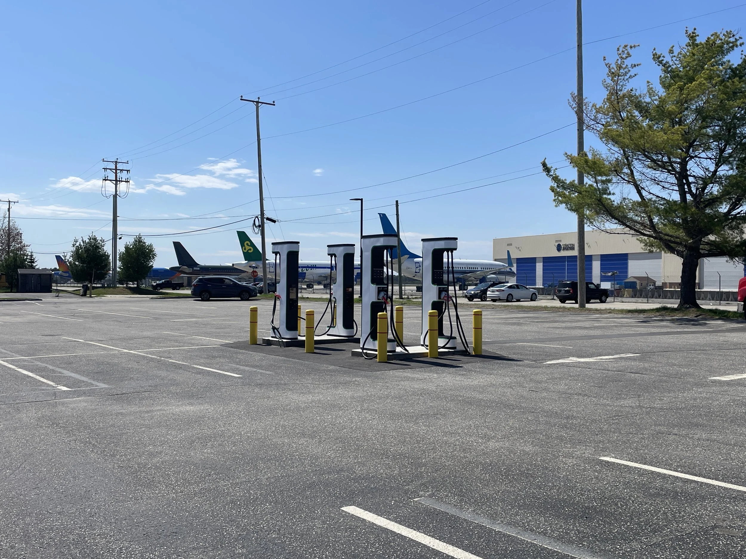 An empty parking lot with six electric vehicle charging stations, parked cars, utility poles, trees, and a large building in the background under a clear blue sky.