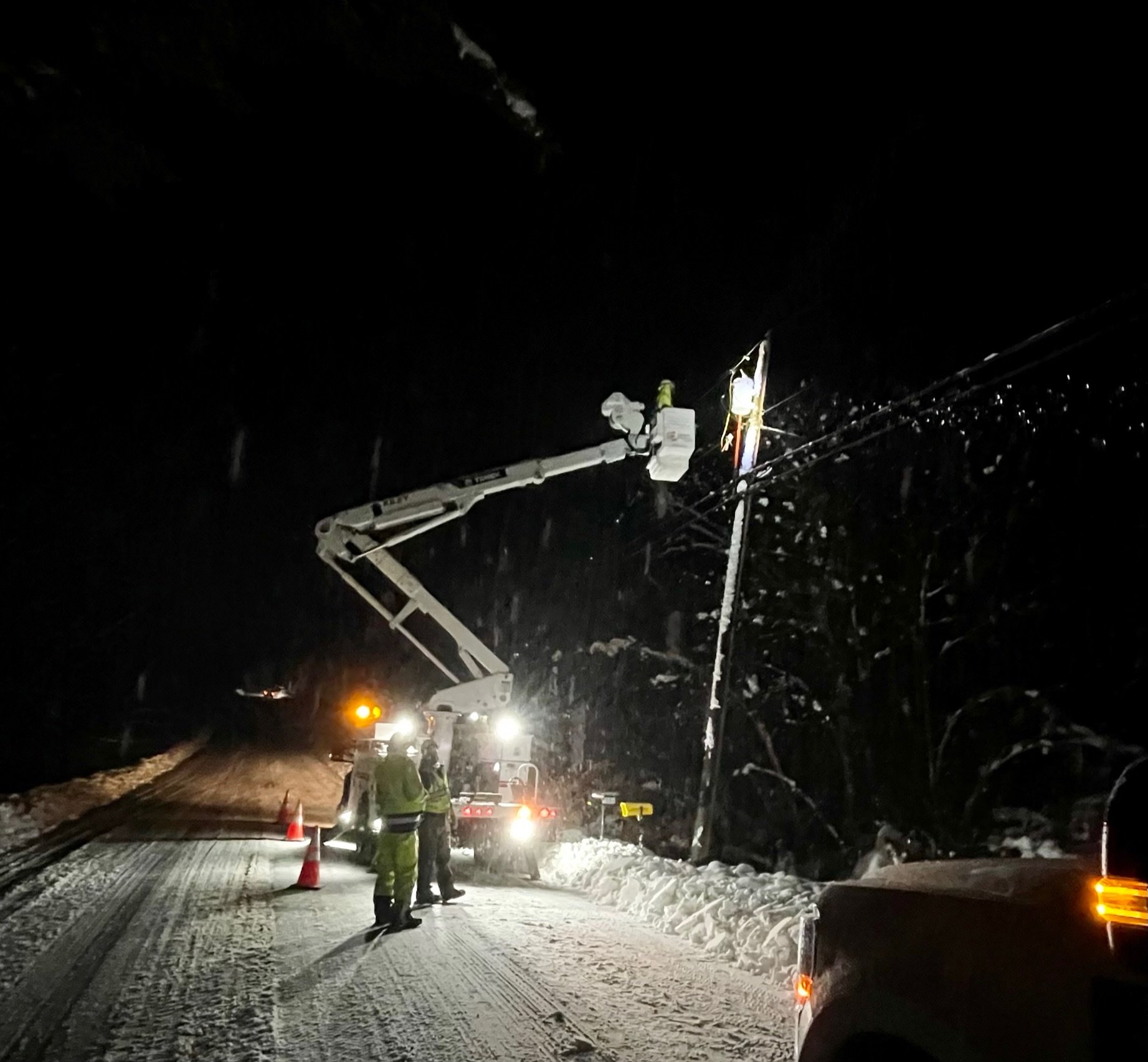 Nighttime scene of utility workers in reflective gear fixing a downed power line. They are using a bucket truck, and street cones are placed around the work area. Snow covers the ground, and the background shows a dark sky and illuminated utility pol
