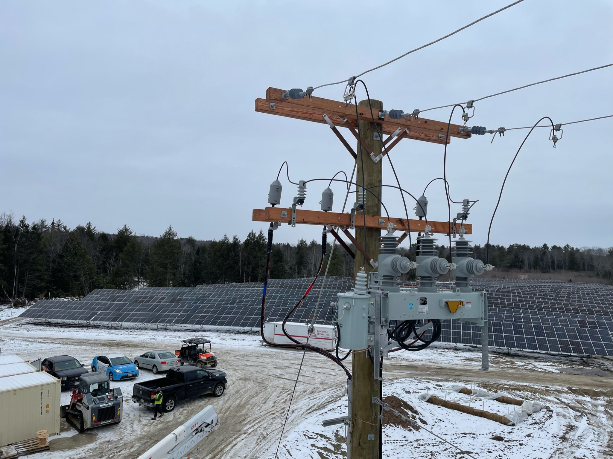 View of a utility pole with electrical wires, solar panels on the ground, and several parked vehicles on snow-covered ground with a forested background under a cloudy sky.