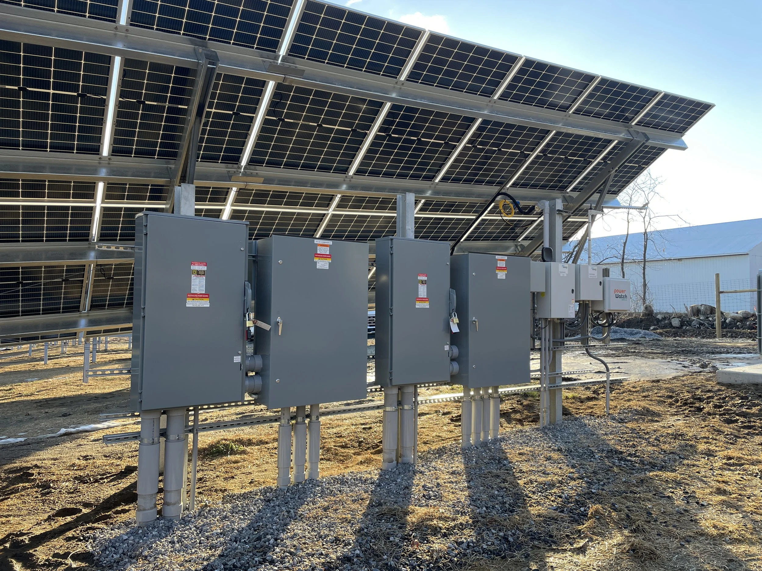 Solar panels on metal structures with electrical boxes and wiring outdoors under a clear blue sky.