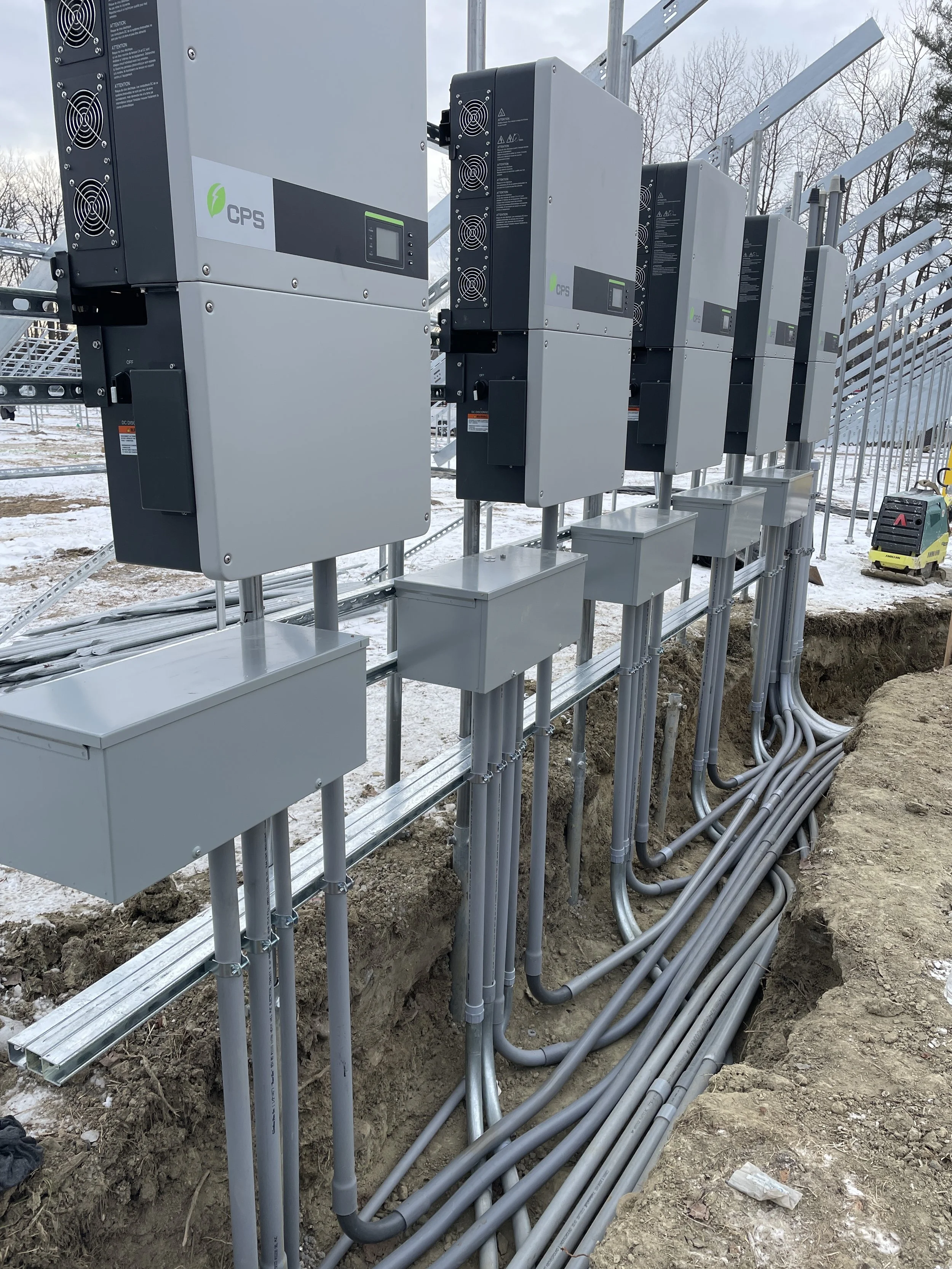 A row of inverters and electrical boxes installed at an outdoor power plant site, with numerous cables running underground and snow on the ground.