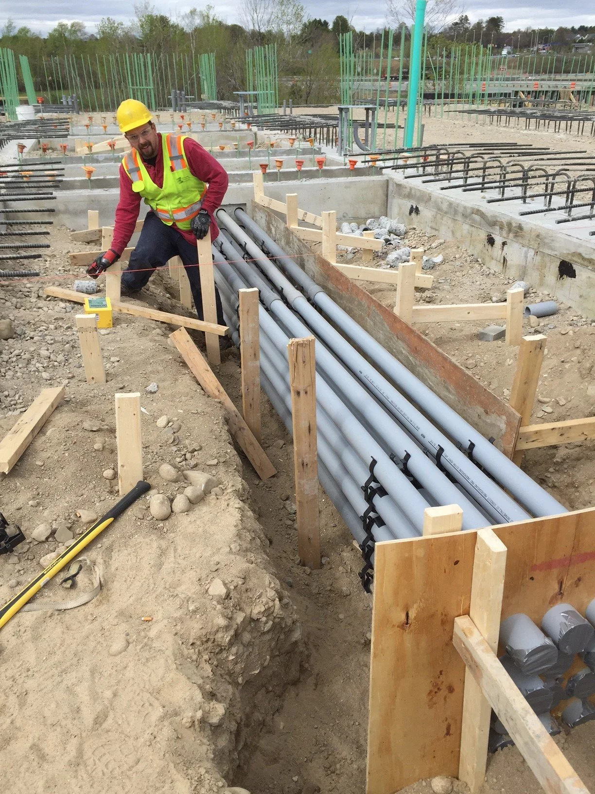 A construction worker with a yellow hard hat and safety vest working at a construction site, installing or inspecting pipes in a trench surrounded by wooden framing, with construction equipment and materials visible in the background.