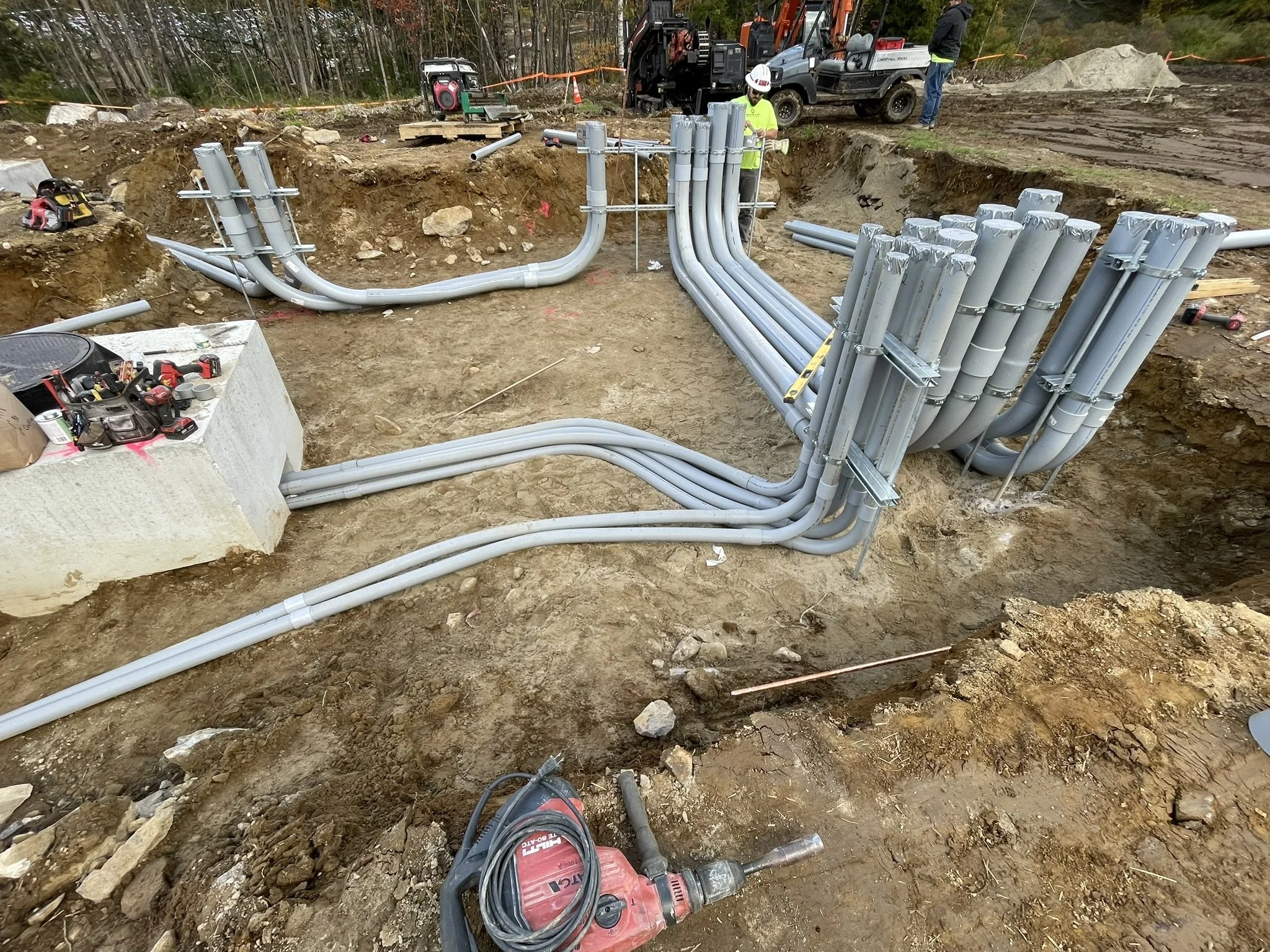 Construction workers installing electrical or utility conduits in a dug-out outdoor area. Construction tools and equipment are visible on site, with trees in the background.